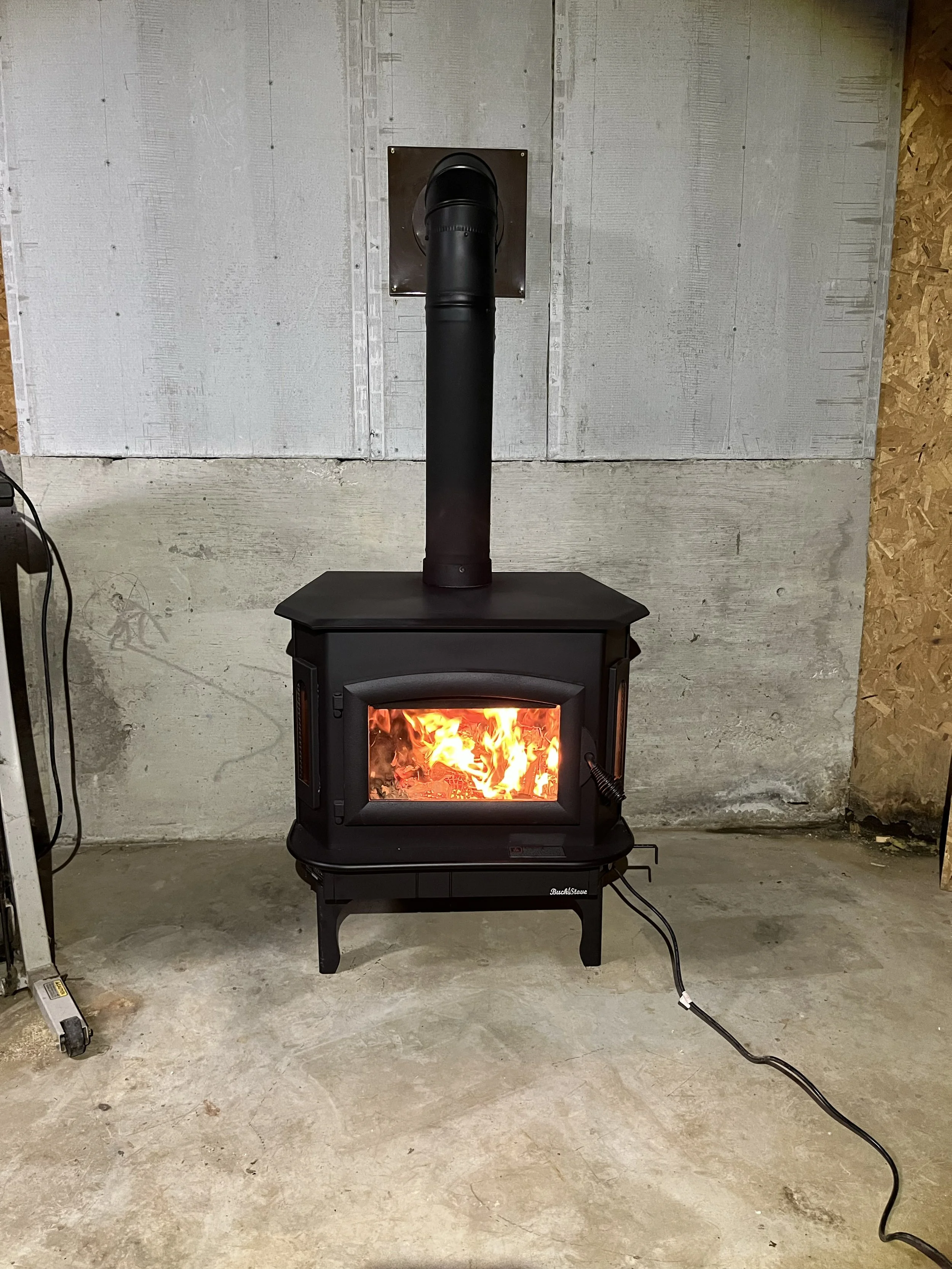 A wood stove with a fire inside, positioned on a black stand, with a black stovepipe extending upward through a square opening in an unfinished wall.