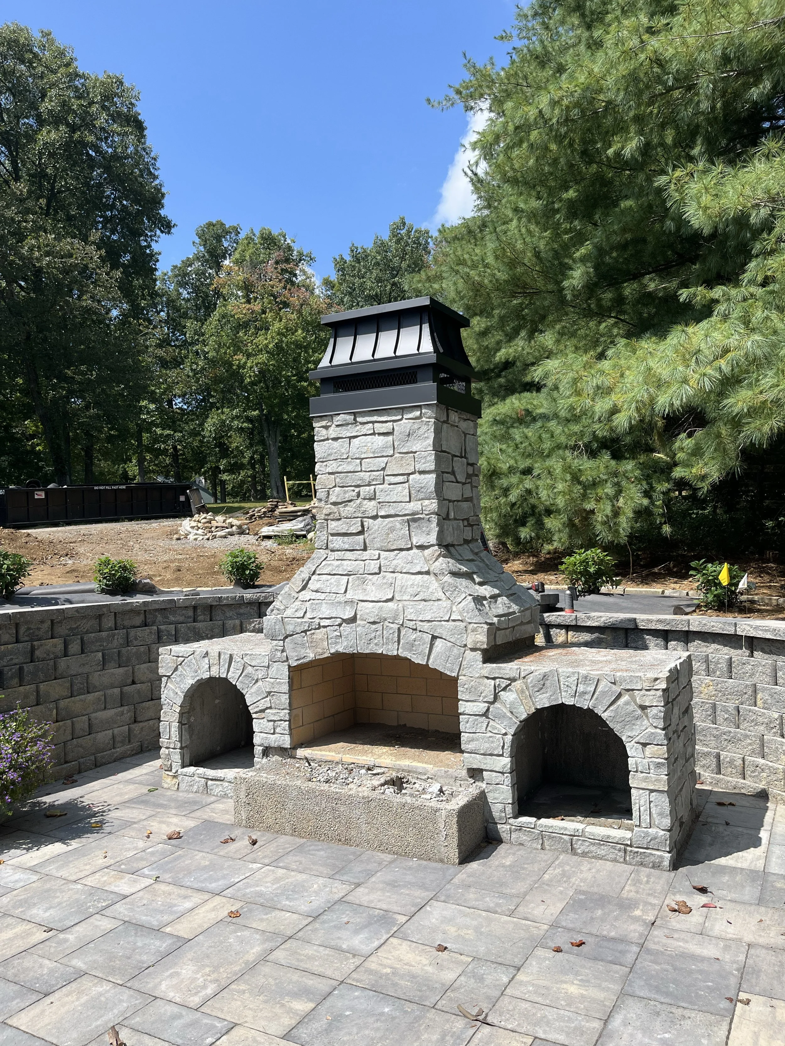 An outdoor stone fireplace with a chimney under construction, surrounded by paving stones and landscaping, with trees and a blue sky in the background.