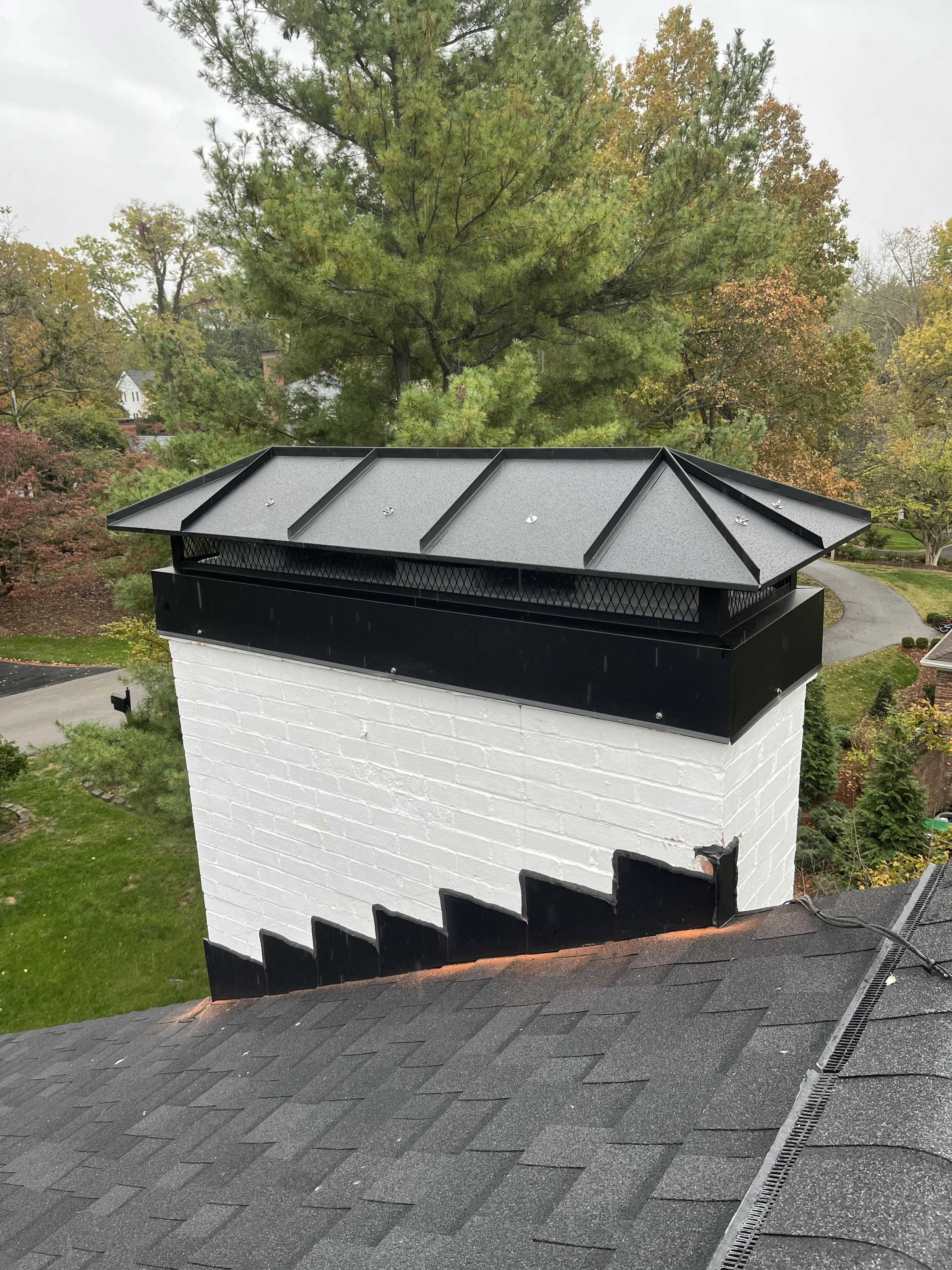View of a white brick chimney with a black metal cap on a sloped roof, surrounded by green and autumn-colored trees.