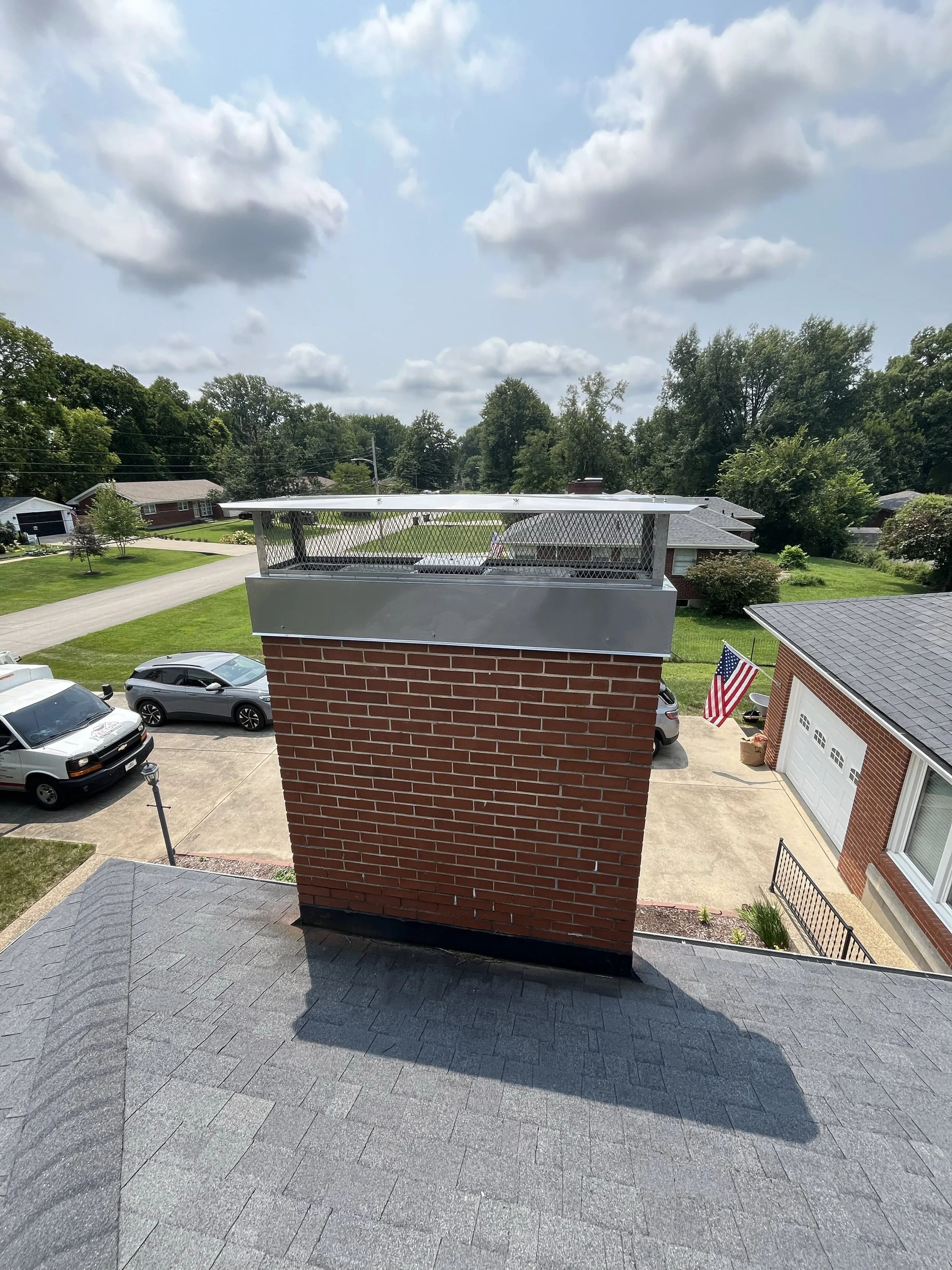 View of a suburban neighborhood from a rooftop, showing a red brick chimney in the foreground, multiple houses, trees, a driveway with parked cars, an American flag, and a partly cloudy sky.