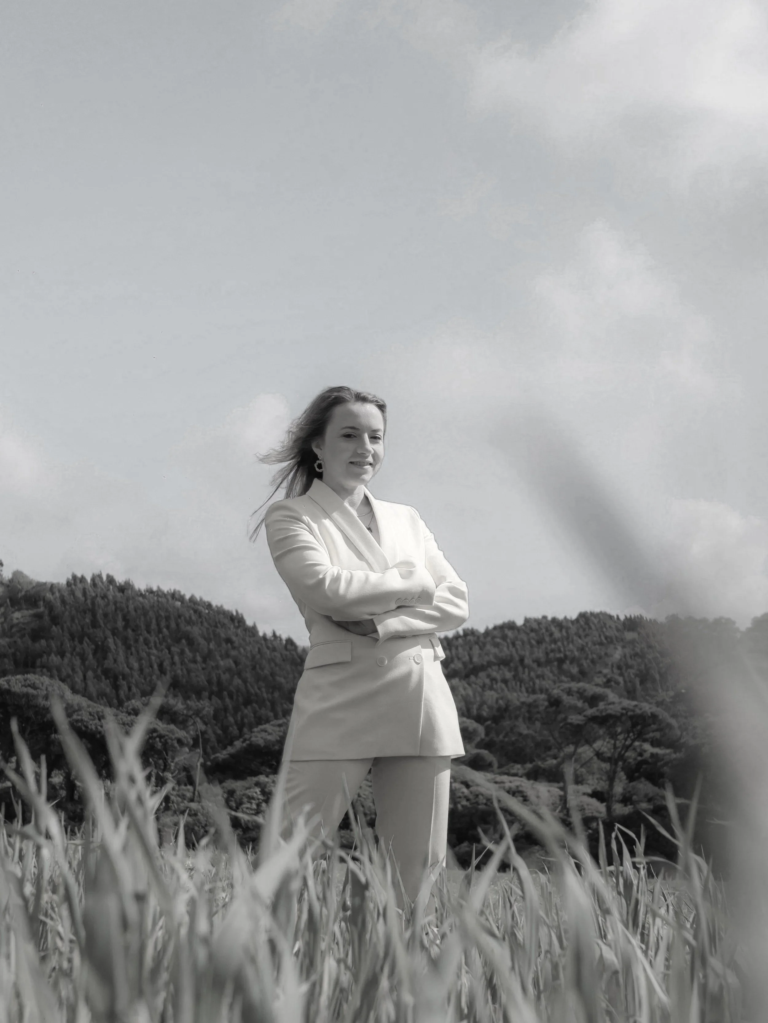 A woman in a suit stands outdoors in a field with mountains and cloudy sky in the background, arms crossed, smiling.