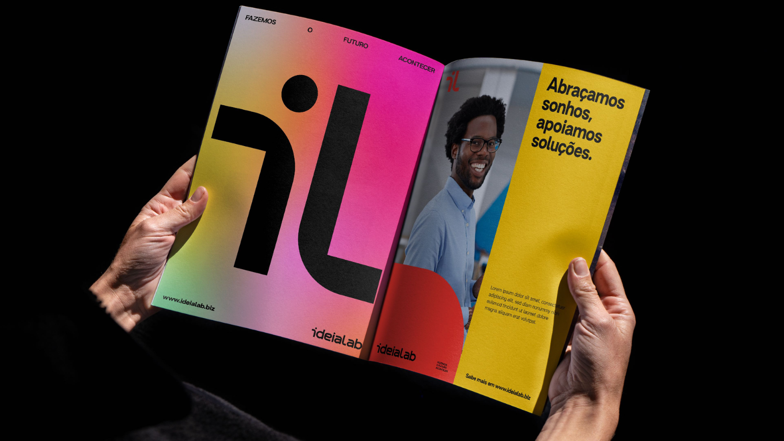 A person holding an open magazine with colorful pages; the left page has a large stylized letter 'i' and the right page features a portrait of a smiling man with dark curly hair, glasses, and a light blue shirt along with Portuguese text on a yellow background.