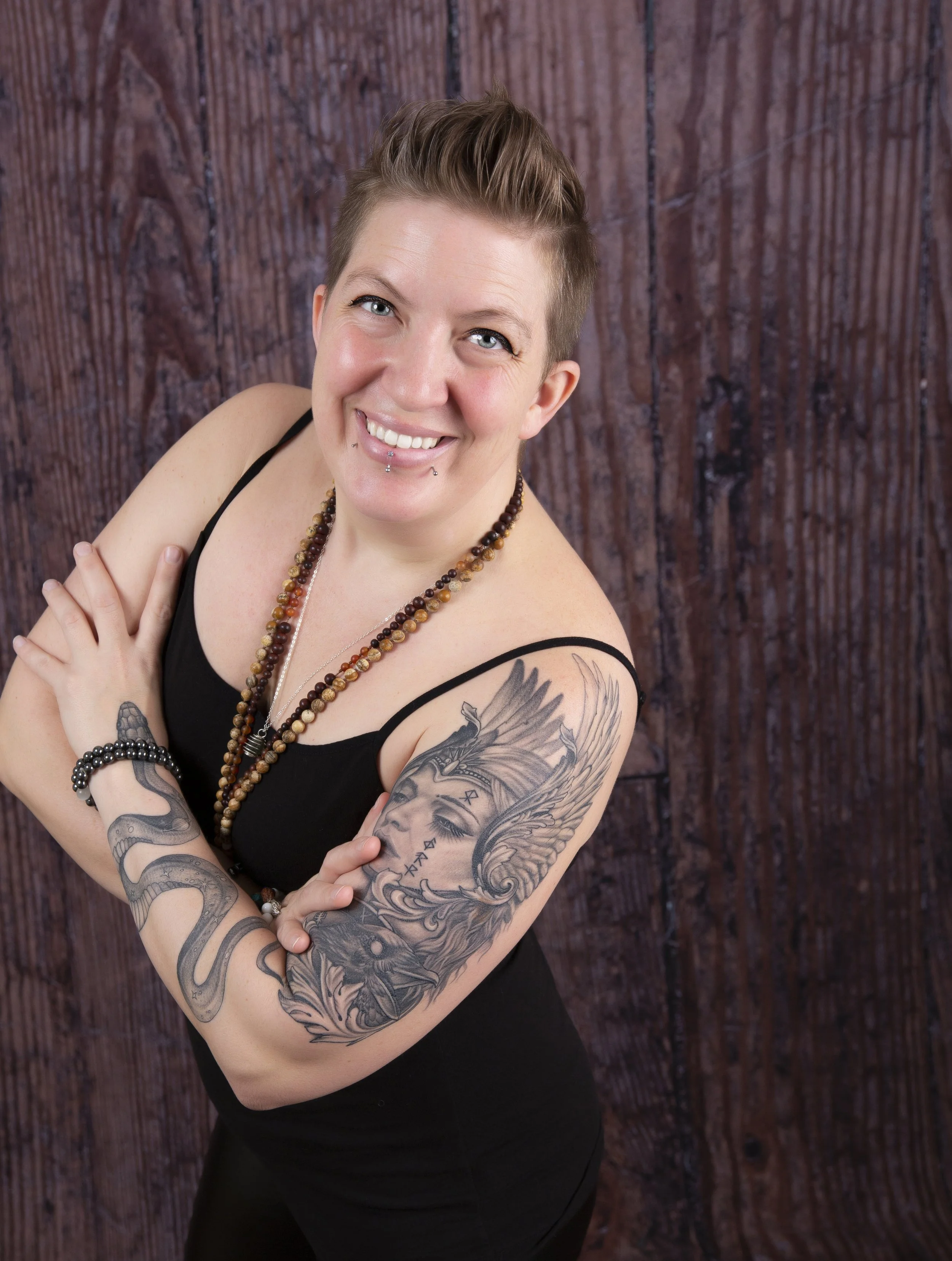 A woman with short hair and tattoos on her arms, wearing a black sleeveless top and beaded necklaces, smiling and standing with her arms crossed in front of a wooden background.