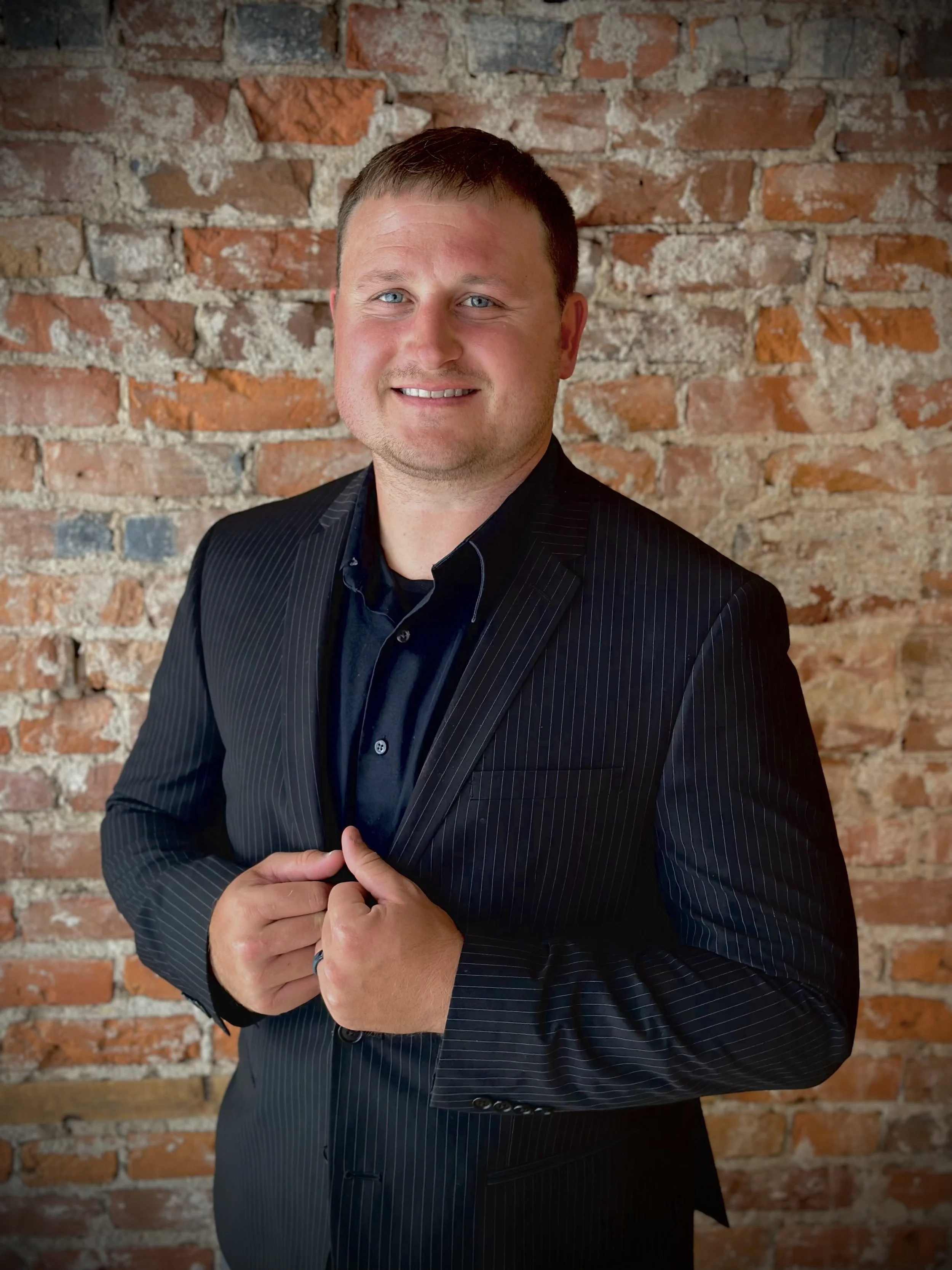A man with short brown hair and blue eyes in a black pinstripe suit, standing in front of a brick wall, smiling and holding his hands together in front of him.
