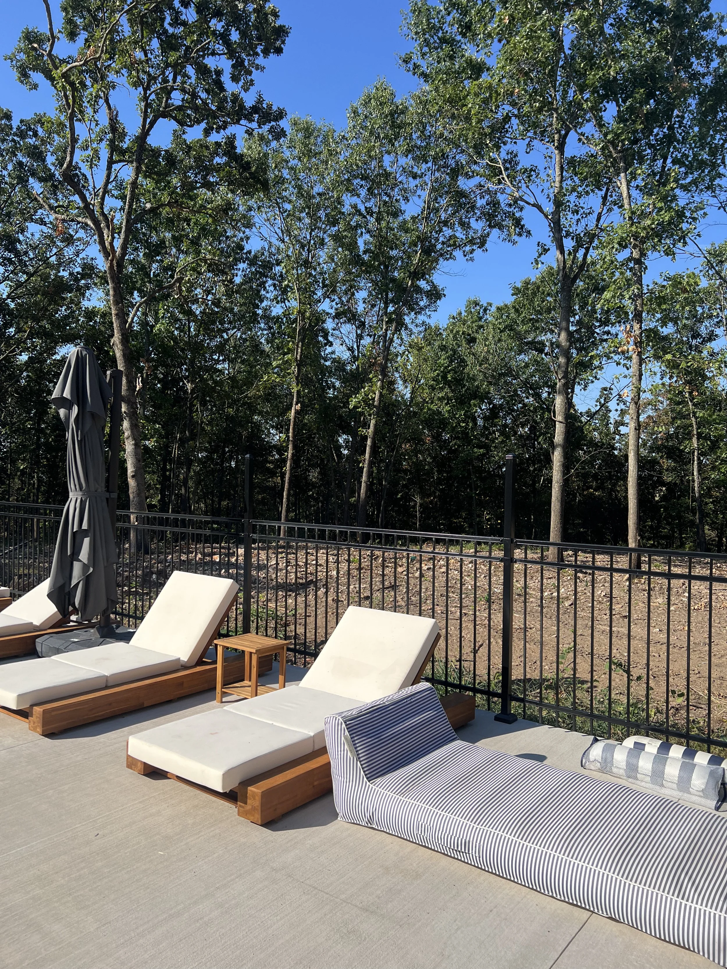 Poolside lounge area with white cushioned sunbeds, a striped cushioned lounge, patio umbrella, small wooden table, black metal fence, and tall trees with green leaves against a clear blue sky.
