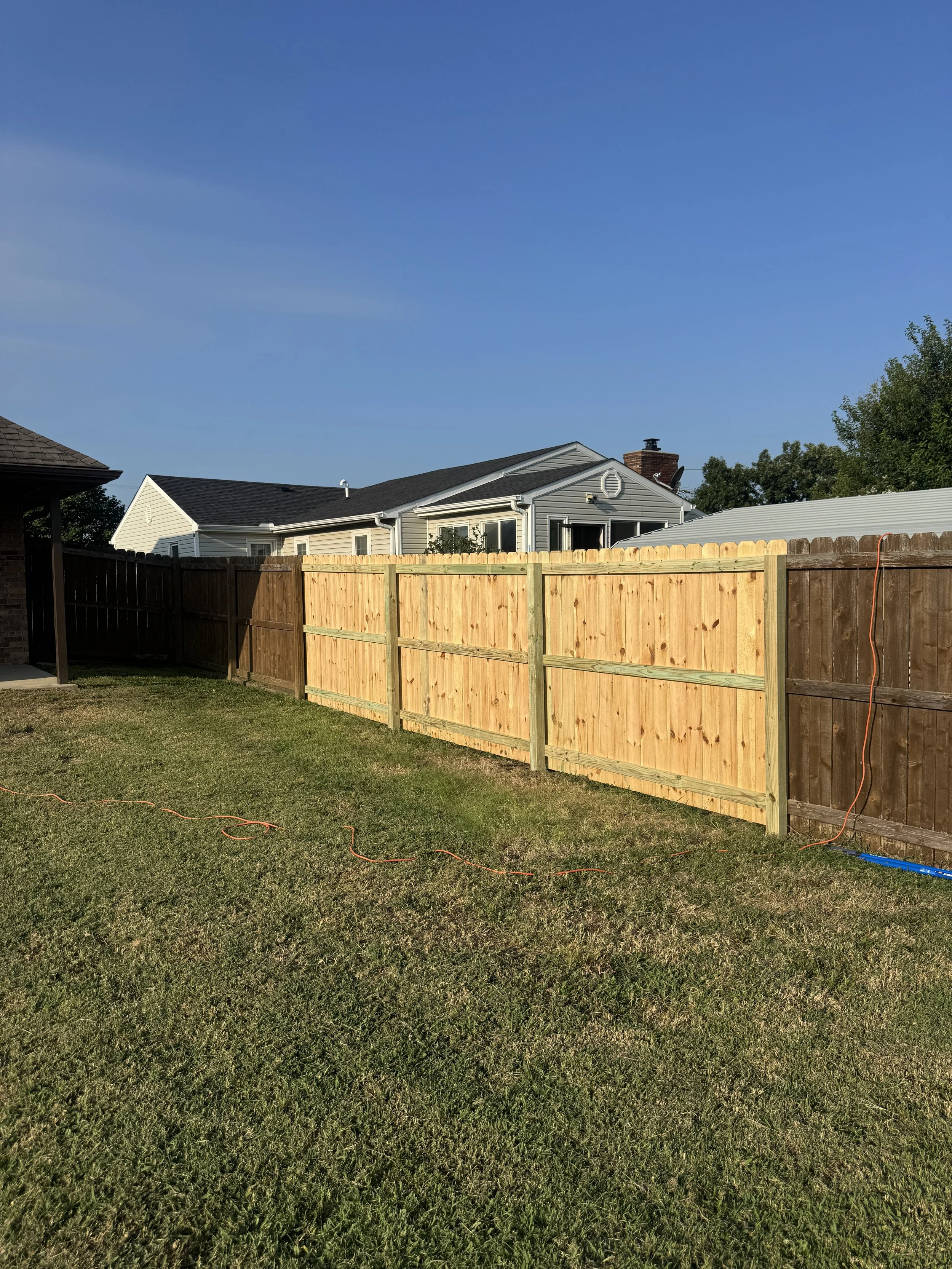 A fenced backyard with grass, a house with a grey roof, a chimney, and a blue sky.