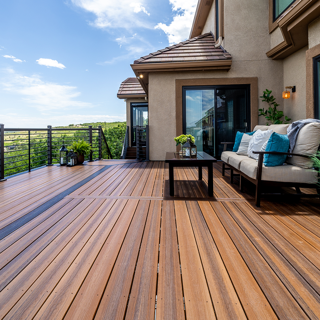 A spacious wooden deck attached to a house with outdoor furniture, potted plants, and a scenic view of greenery and a blue sky with some clouds.