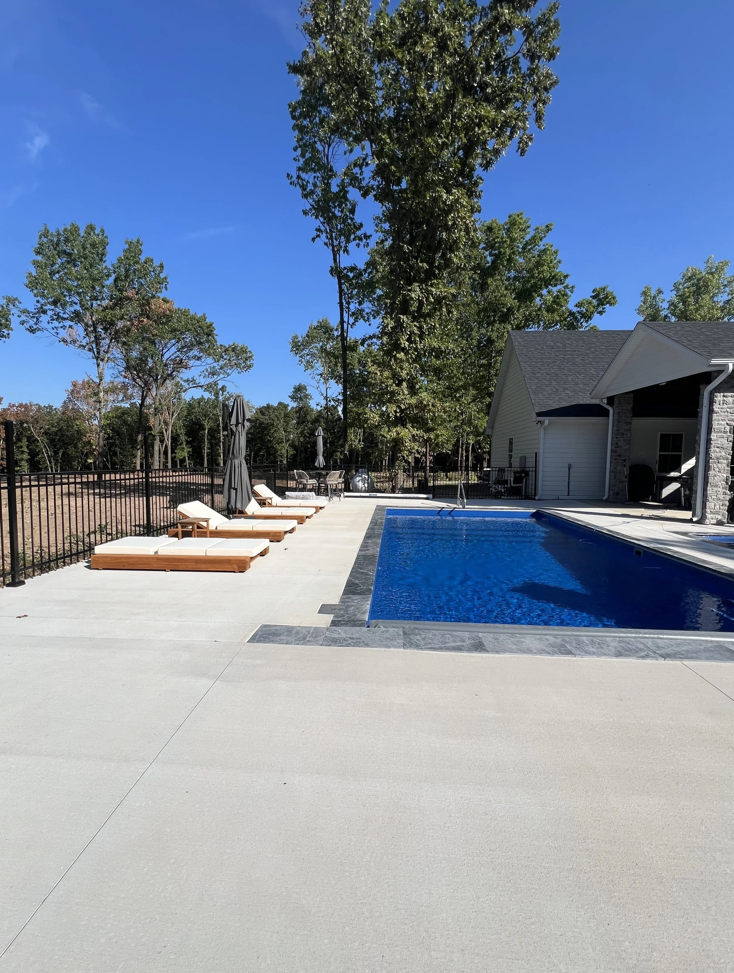 An outdoor swimming pool area with lounge chairs, umbrellas, a house with stone and siding exterior, and trees in the background under a clear blue sky.