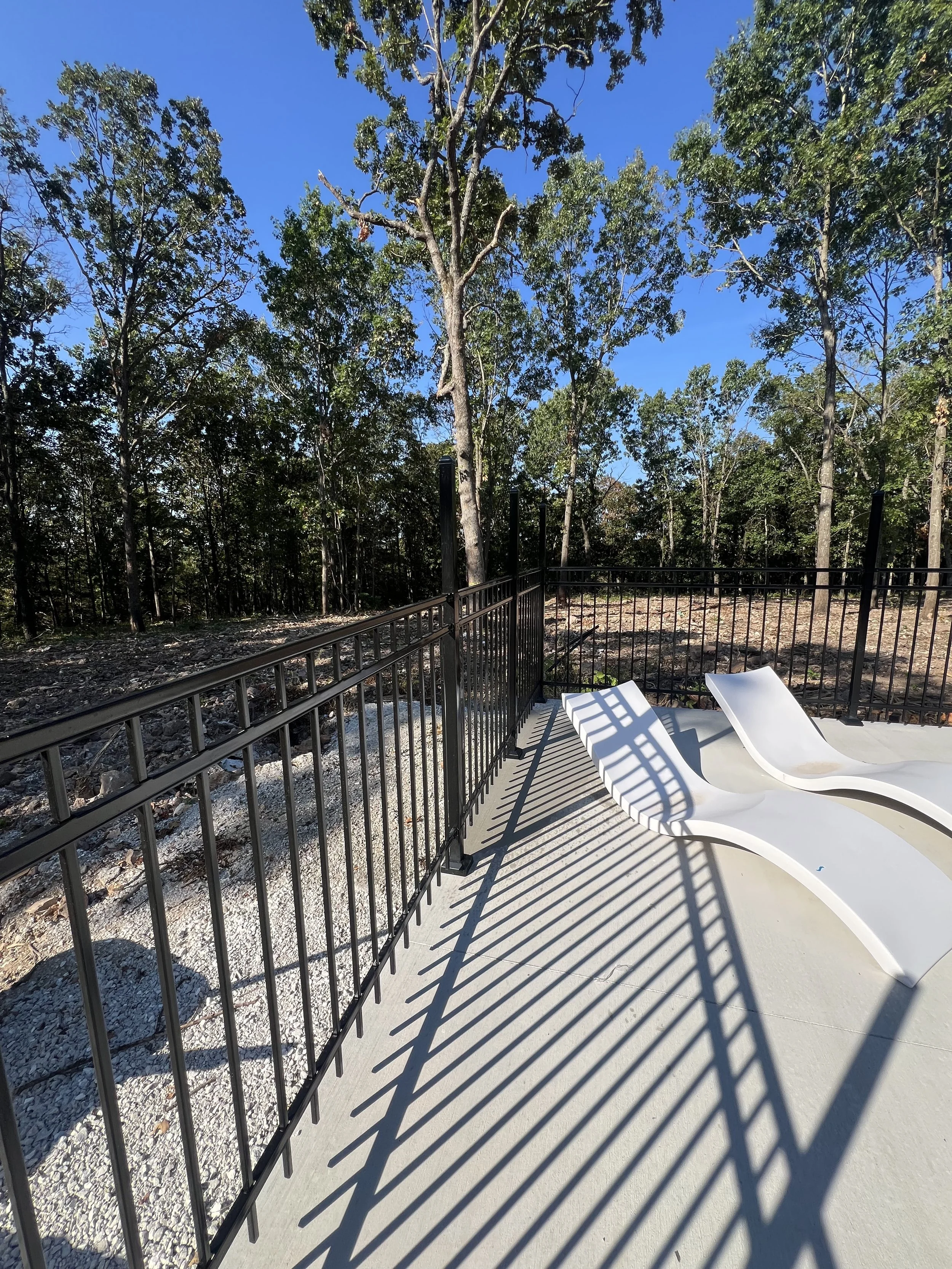 A backyard with a concrete patio and two white lounge chairs, enclosed by black metal fencing, overlooking a wooded area with trees and blue sky.