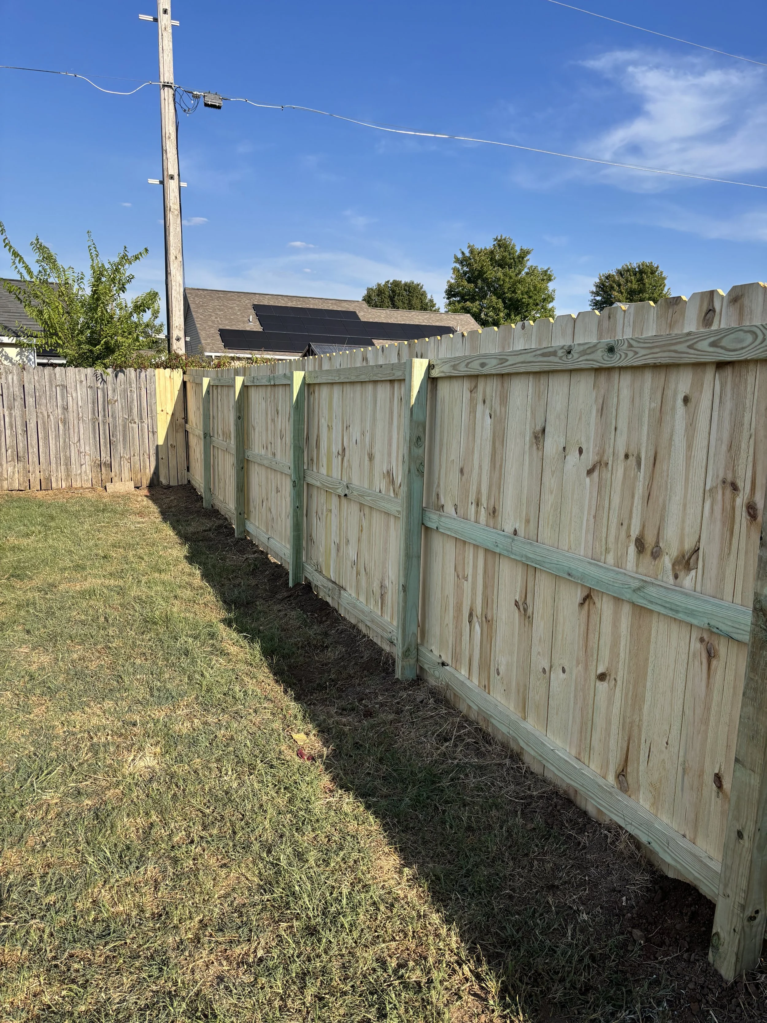 A backyard scene with a newly constructed wooden fence, a strip of grass, and a clear blue sky with some clouds. In the background, there are houses with solar panels on the roof and an electrical pole with wires.