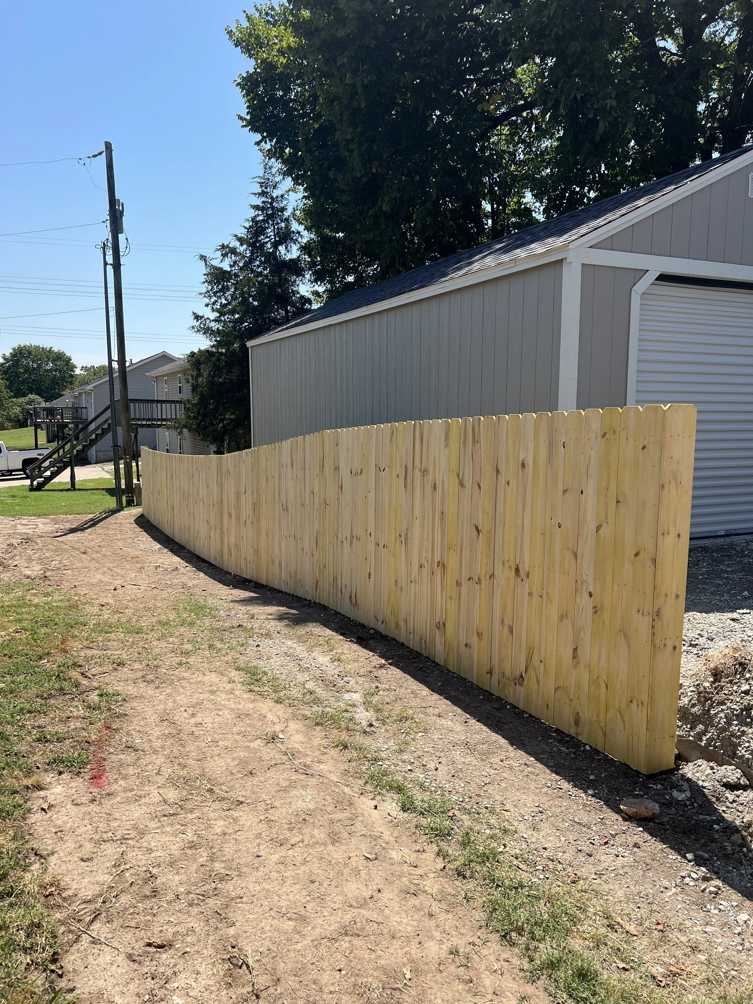 A newly built beige wooden fence runs along a dirt patch in a backyard. In the background, there is a light gray building with vertical siding and a black shingled roof. To the left, an electrical pole with multiple wires stands near a set of stairs attached to a neighboring house with beige siding and multiple decks. There are trees with green leaves behind the buildings, and the sky is clear and blue.