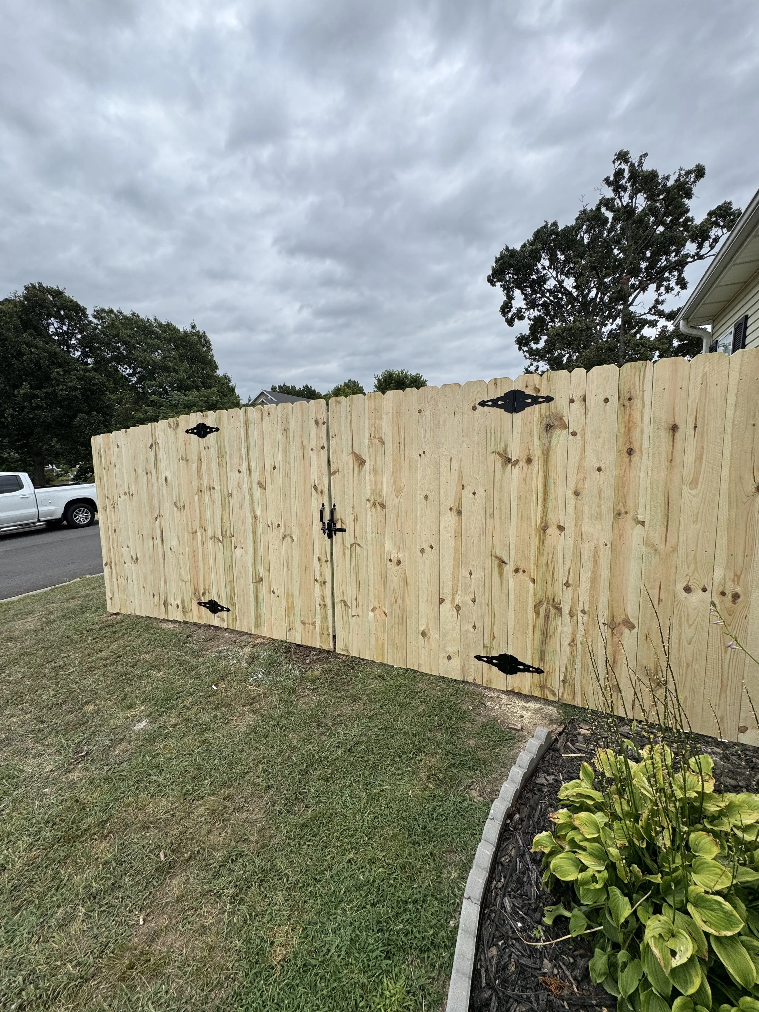 A wooden fence with black decorative hinges and latch in front of a yard with grass, trees, and a partly cloudy sky.