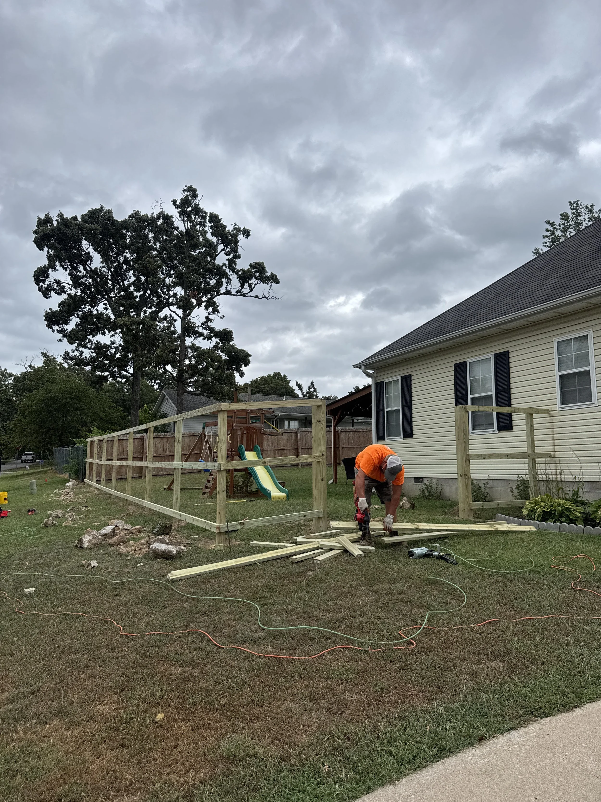 A person working on building a wooden fence in a backyard during overcast weather, with a house, playground slide, and trees visible in the background.