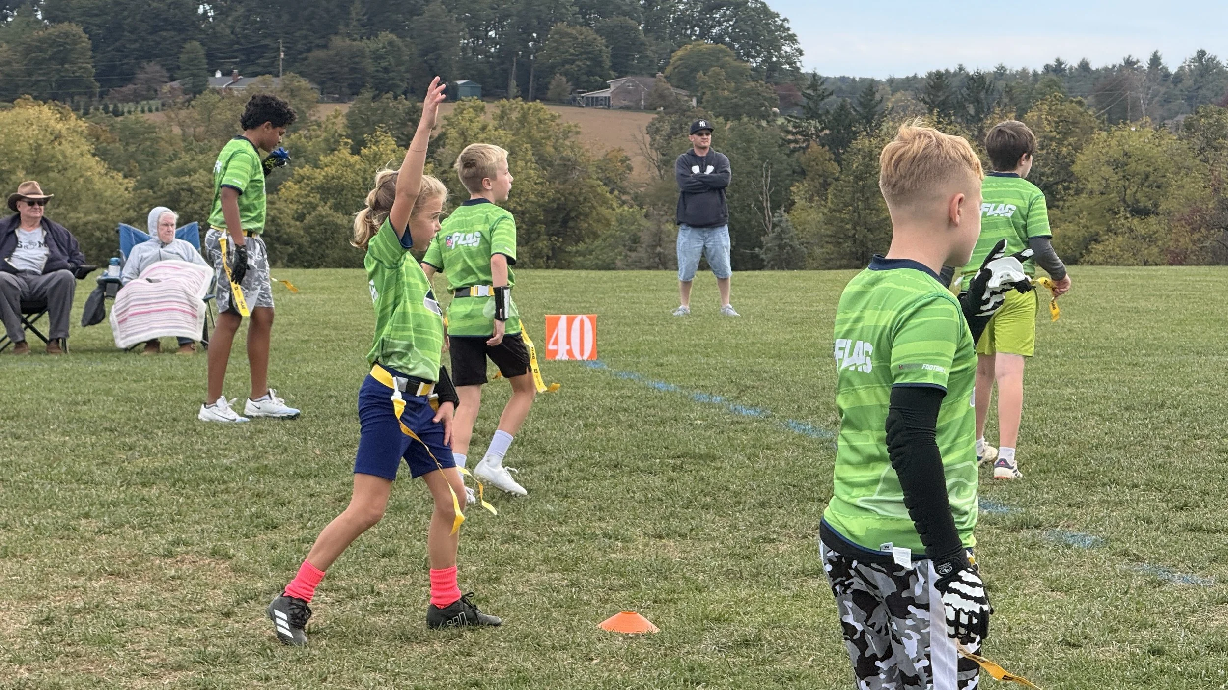 Children in green and black sports uniforms participating in a flag football game on a grassy field, with spectators and trees in the background.