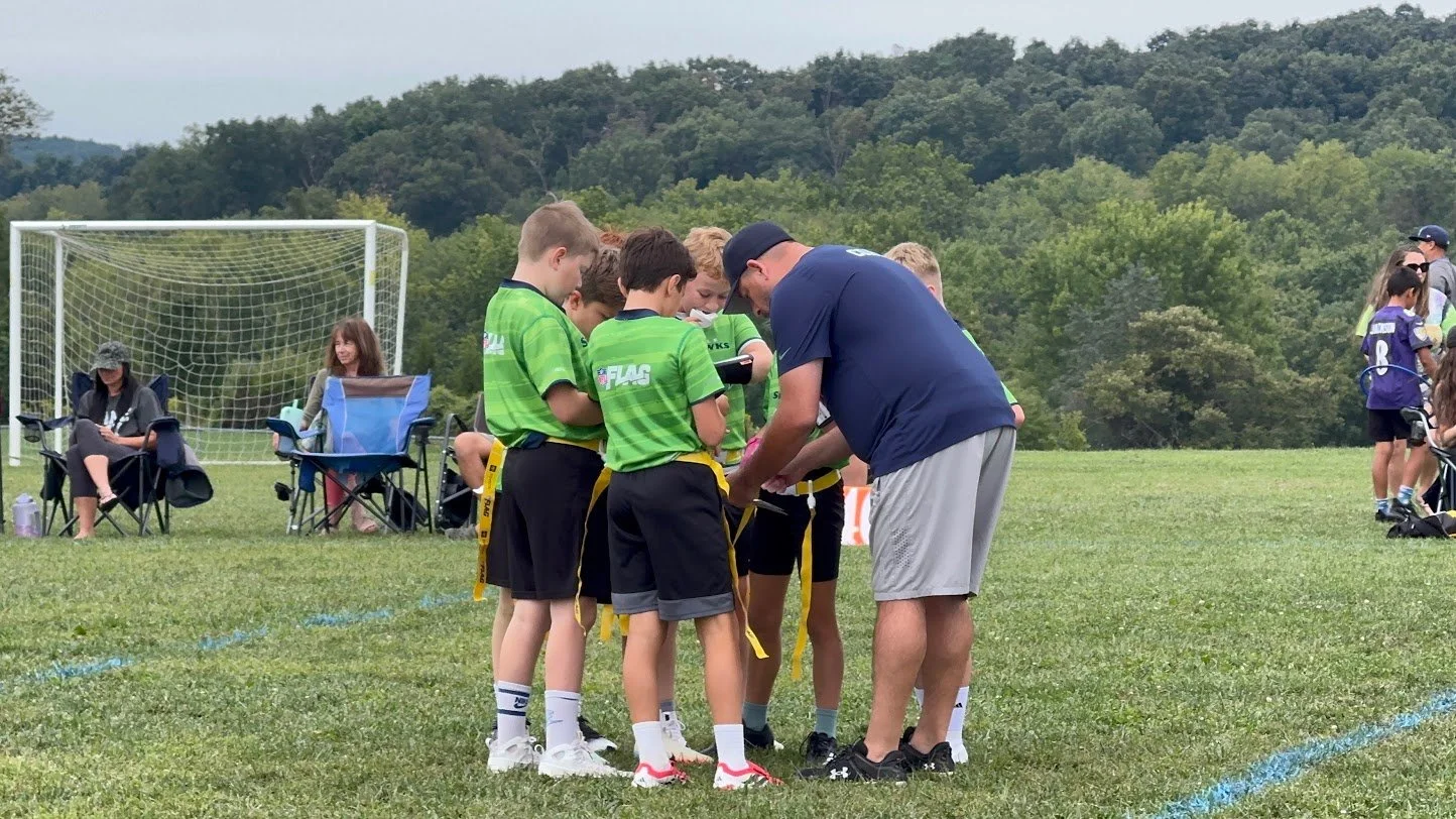 A group of young boys in green soccer jerseys receiving awards from a coach on a grassy field during a soccer event, with spectators sitting in chairs and a goal net in the background.