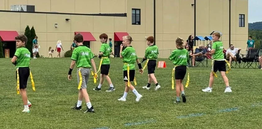 Young boys in green sports jerseys and black shorts walking on a grassy field, possibly during a sports event or game, with spectators seated on the sideline.