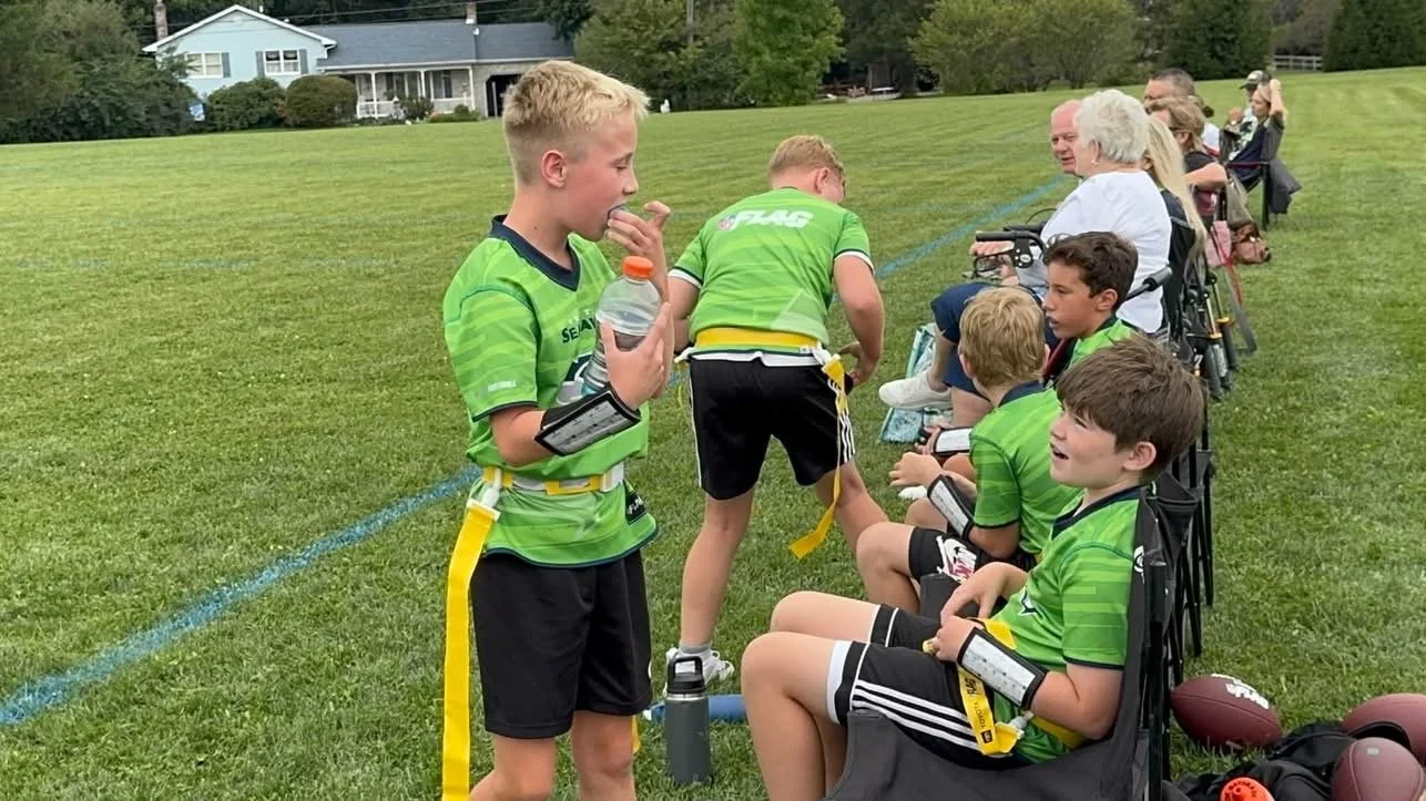 Group of young boys wearing green jersey football uniforms sitting on a bench on a grassy field, with some adults standing behind them, preparing for or after a game.