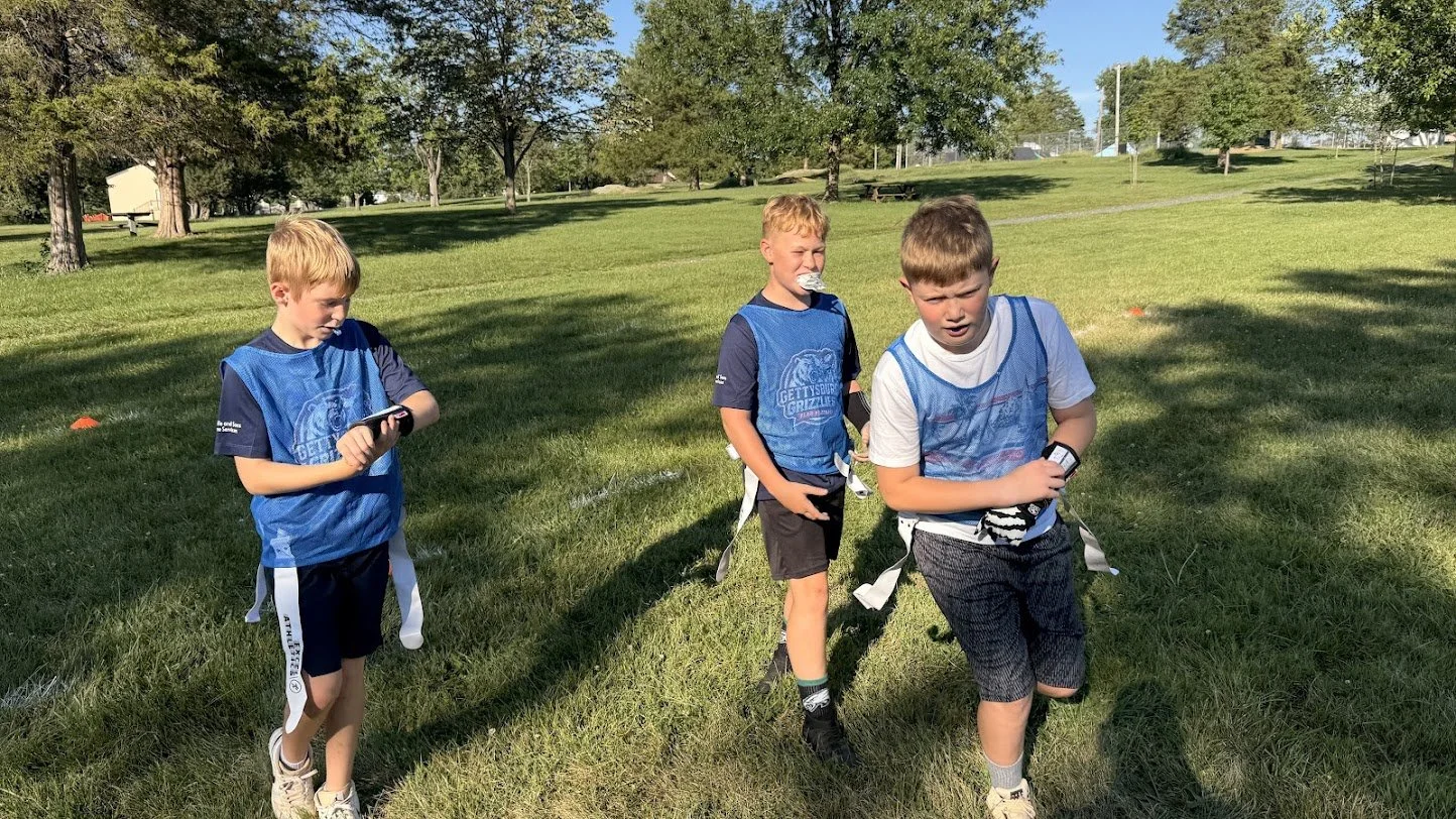 Three young boys in blue sports jerseys walking on a grassy field, with two of them looking at fitness trackers and one with a crumpled paper napkin near his nose. The field is shaded by trees, and there are other trees and a small building in the ba