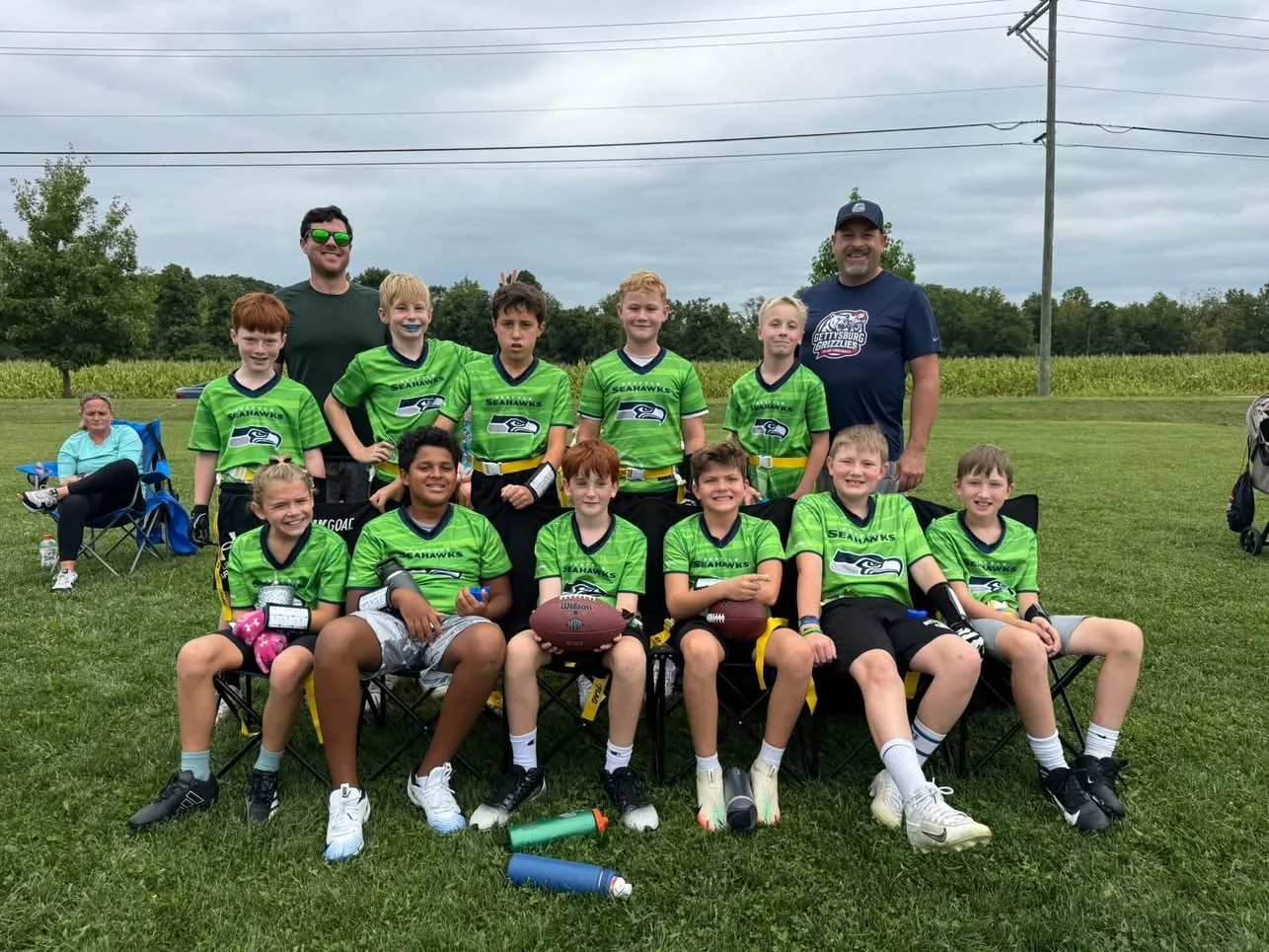 Youth football team in green jerseys with the Seahawks logo, posing with their coaches outdoors on grass, along with a woman sitting in a chair and a water bottle nearby.