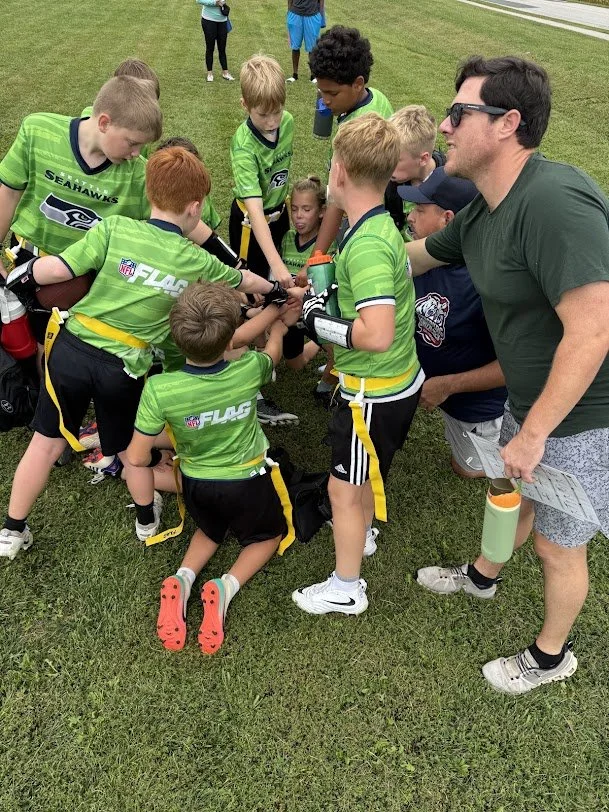 A group of young football players wearing green jerseys huddles together on the grass, receiving instructions from an adult coach during a practice or game.