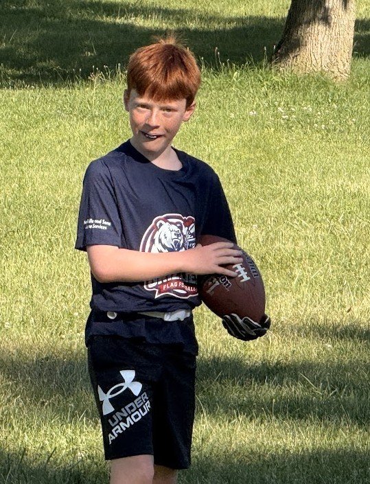 A young boy with red hair wearing a navy blue t-shirt and black shorts holding a football on a grassy field.