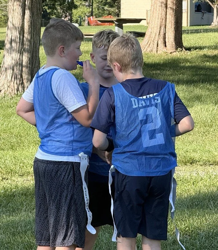 Three young boys in blue jerseys and athletic shorts stand in a circle on a grassy field, having a discussion, with trees and a building in the background.