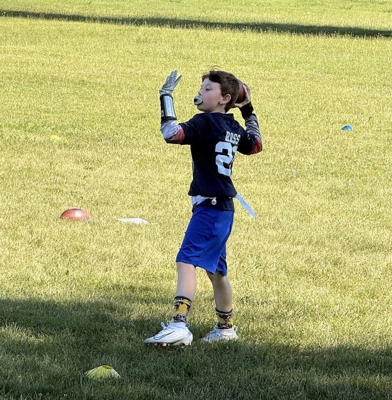 A young boy standing on a grassy field with sports equipment, wearing a football jersey, shorts, sneakers, and a prosthetic arm, with a pacifier in his mouth and surrounded by colorful cones.