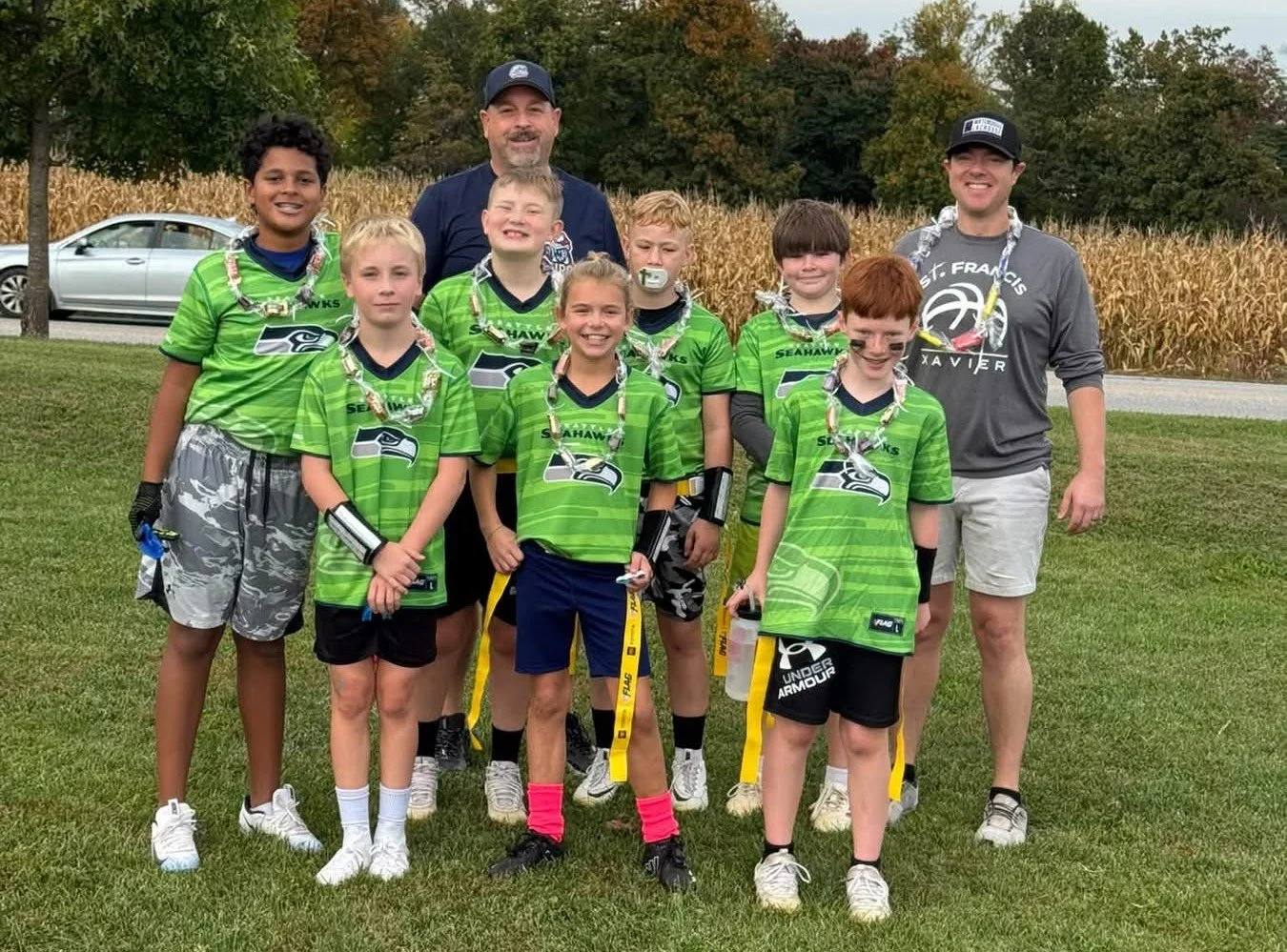 Youth football team in green Jerseys with two adult coaches on a grassy field, some kids with face paint, a silver car in background, and autumn trees.