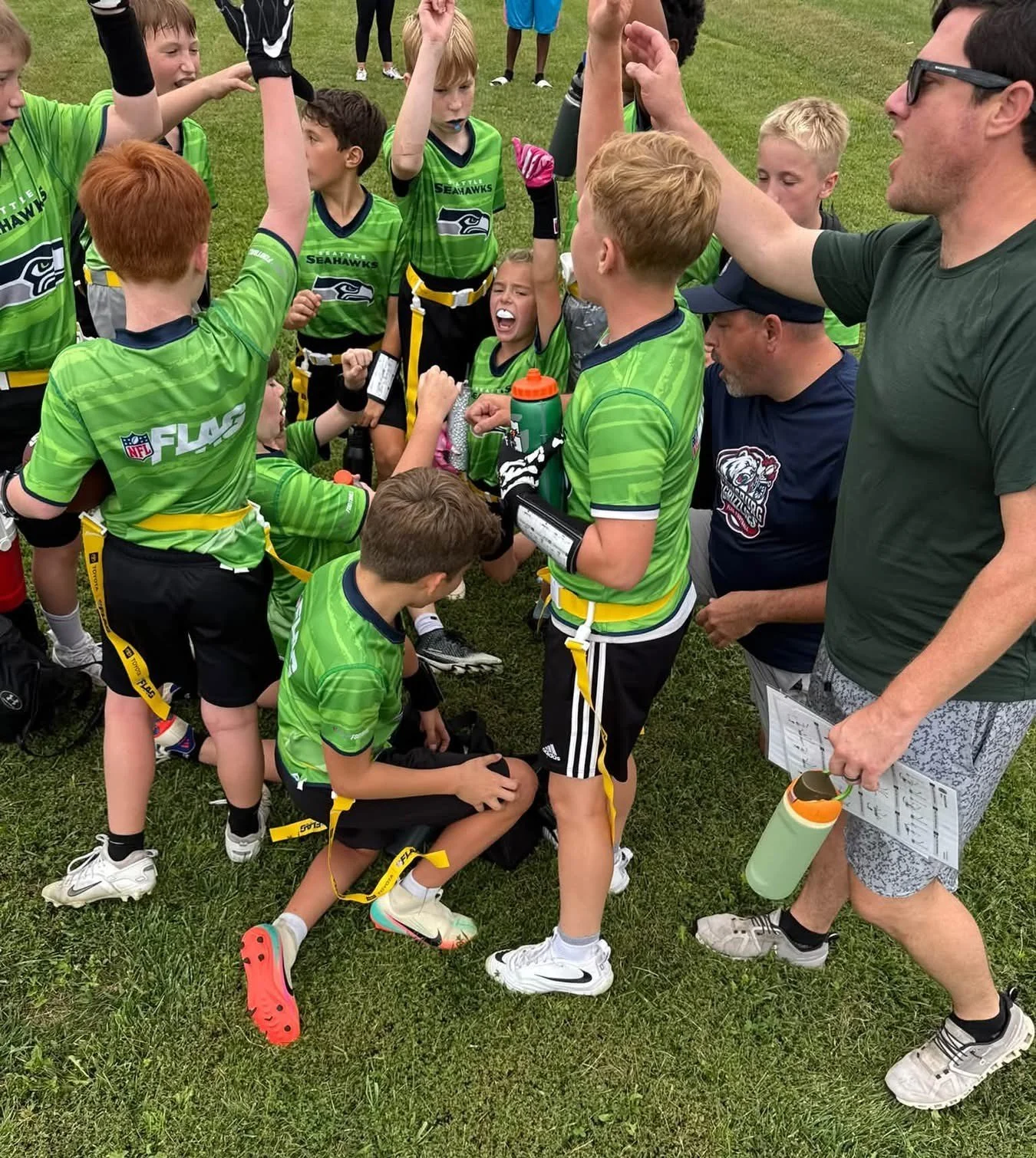 Youth football team huddled with coach on the field, celebrating a game, wearing bright green jerseys with the Seahawks logo.
