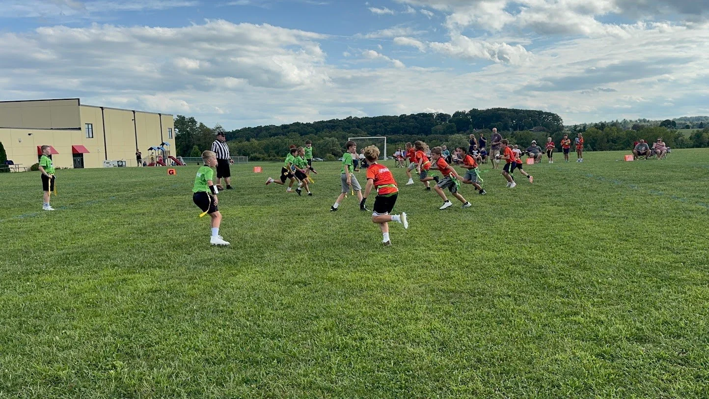 Children playing flag football on a grassy field with adults and a building in the background during daytime.