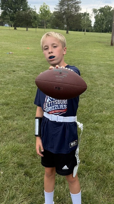 A young boy in a football uniform standing outdoors on a grassy field, holding a football towards the camera with his right hand, with a blue mouthguard in his mouth.