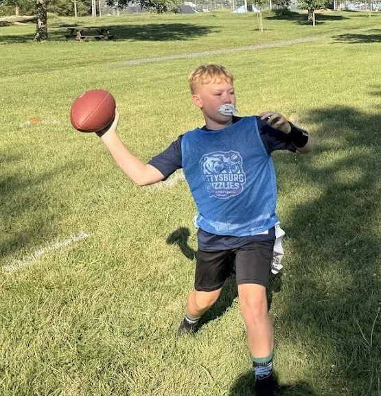Young boy with blond hair, wearing a blue football jersey, running on grass with a football in his right hand and a whistle in his mouth.