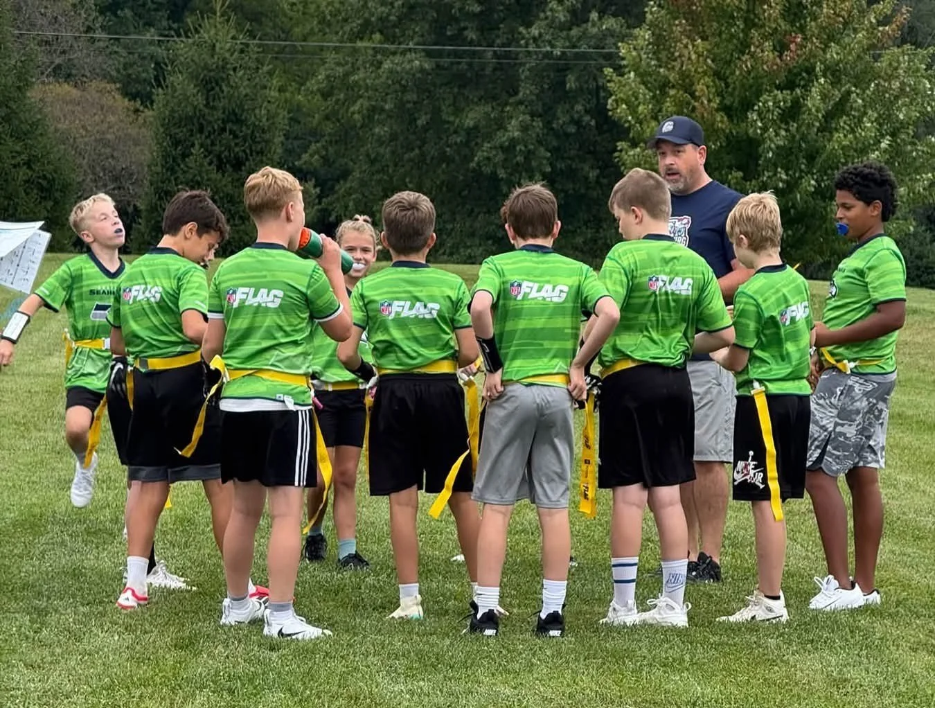 Youth football team in green jerseys on a field listening to coach during practice or game.