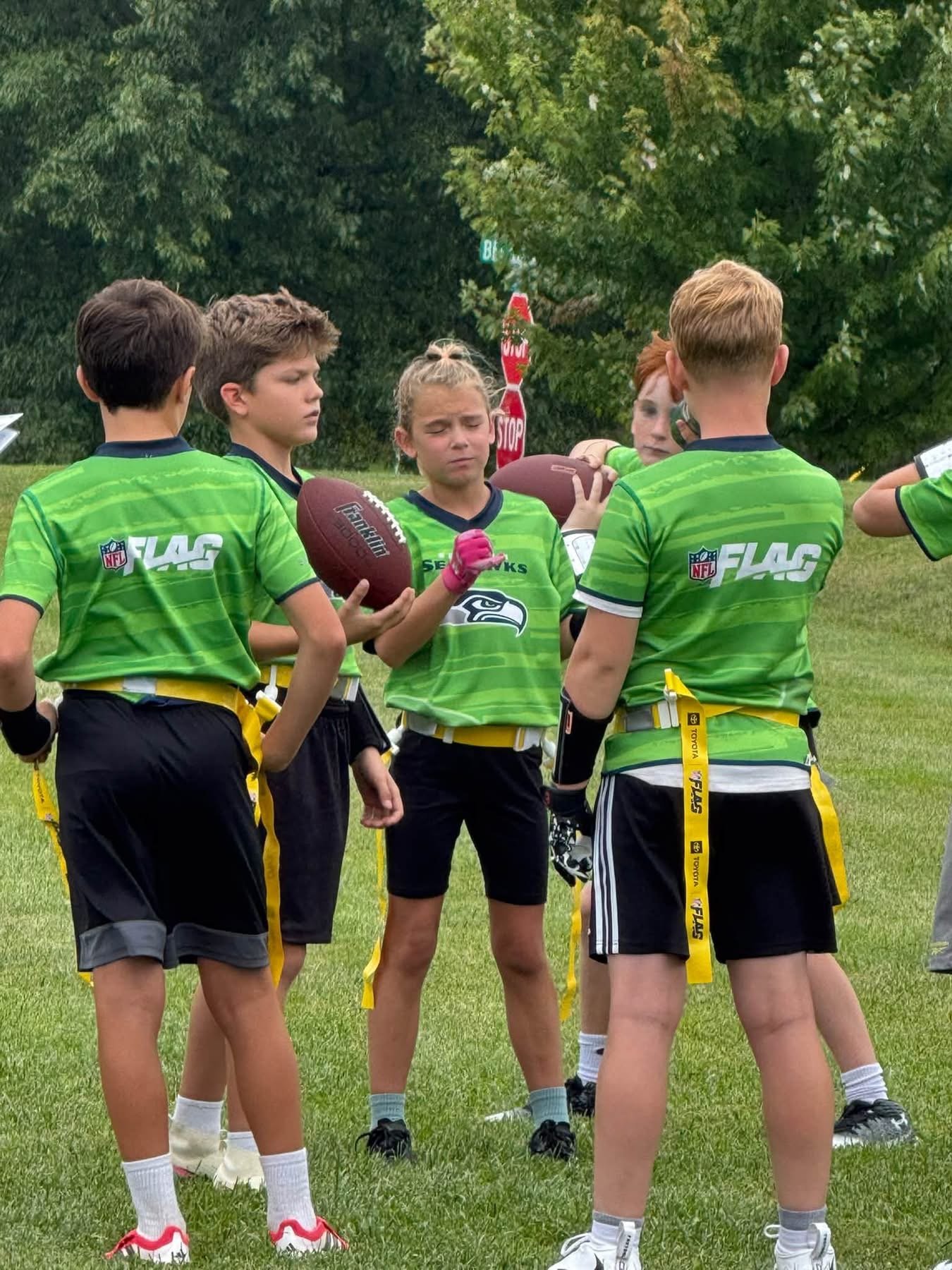 Group of young football players in green jerseys standing in a circle on a grassy field during a game or practice, with trees in the background.