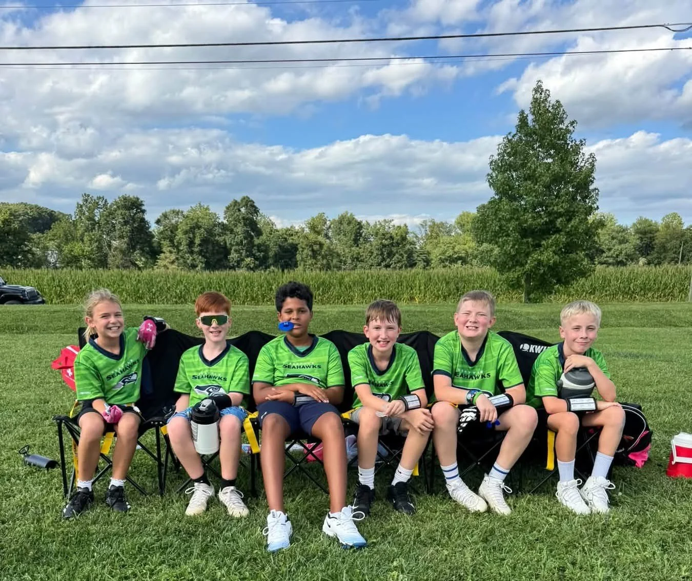 Group of six young boys and girls in green football jerseys, sitting on foldable chairs on a grassy field, smiling with trees and a cloudy sky in the background.