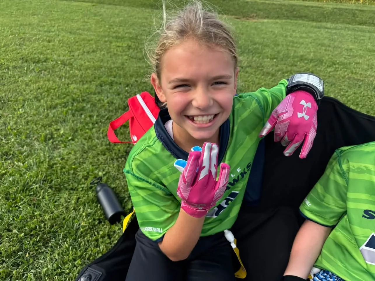 Young girl in a green sports jersey and pink gloves smiling and making a peace sign while sitting on grass with a red backpack behind her.