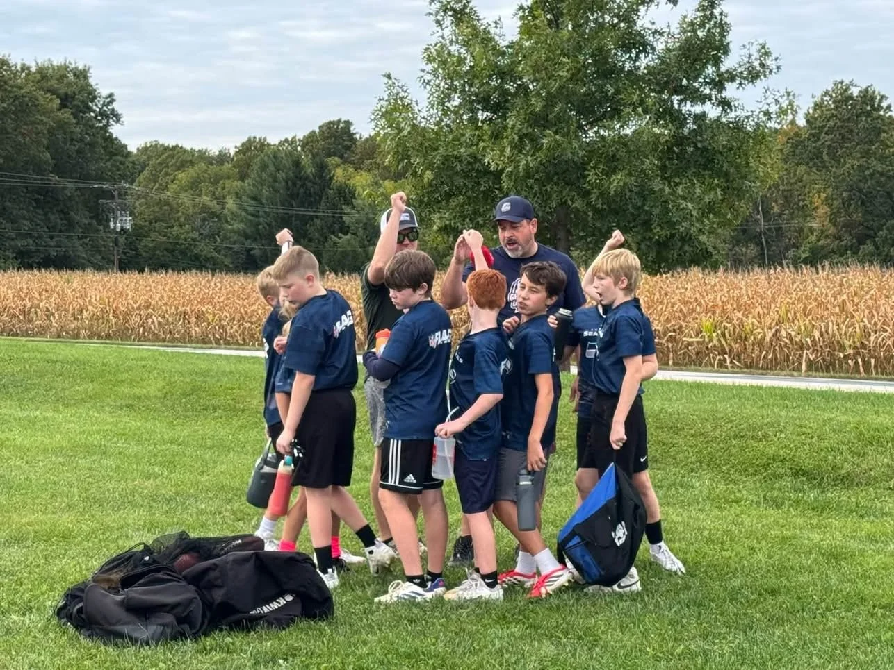 A youth football team huddles with two coaches on a grassy field, with trees and a cornfield in the background.