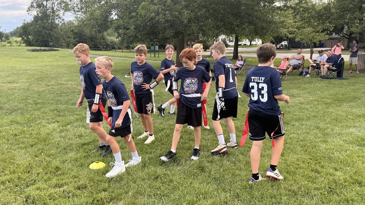Young boys in field hockey uniforms standing on grass, some holding red flags, with adults sitting nearby on lounge chairs watching.