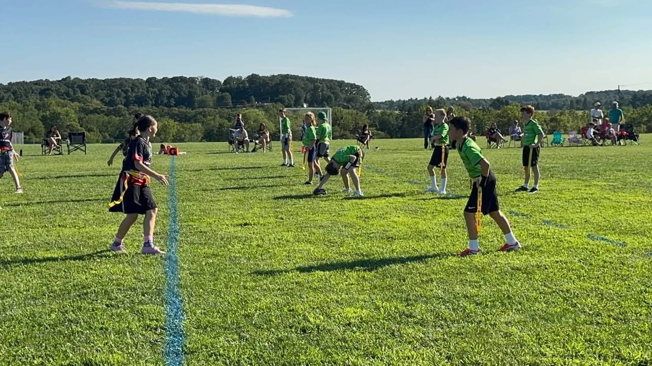Children playing flag football on a grassy field with spectators sitting on chairs in the background.