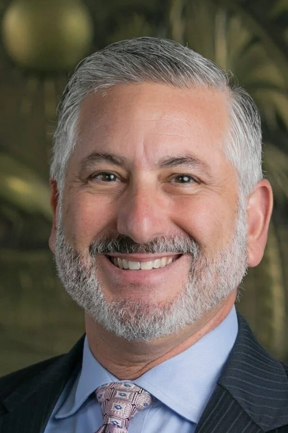 A smiling middle-aged man with gray hair and a beard, wearing a dark suit, light blue shirt, and patterned tie, with a blurred green background.