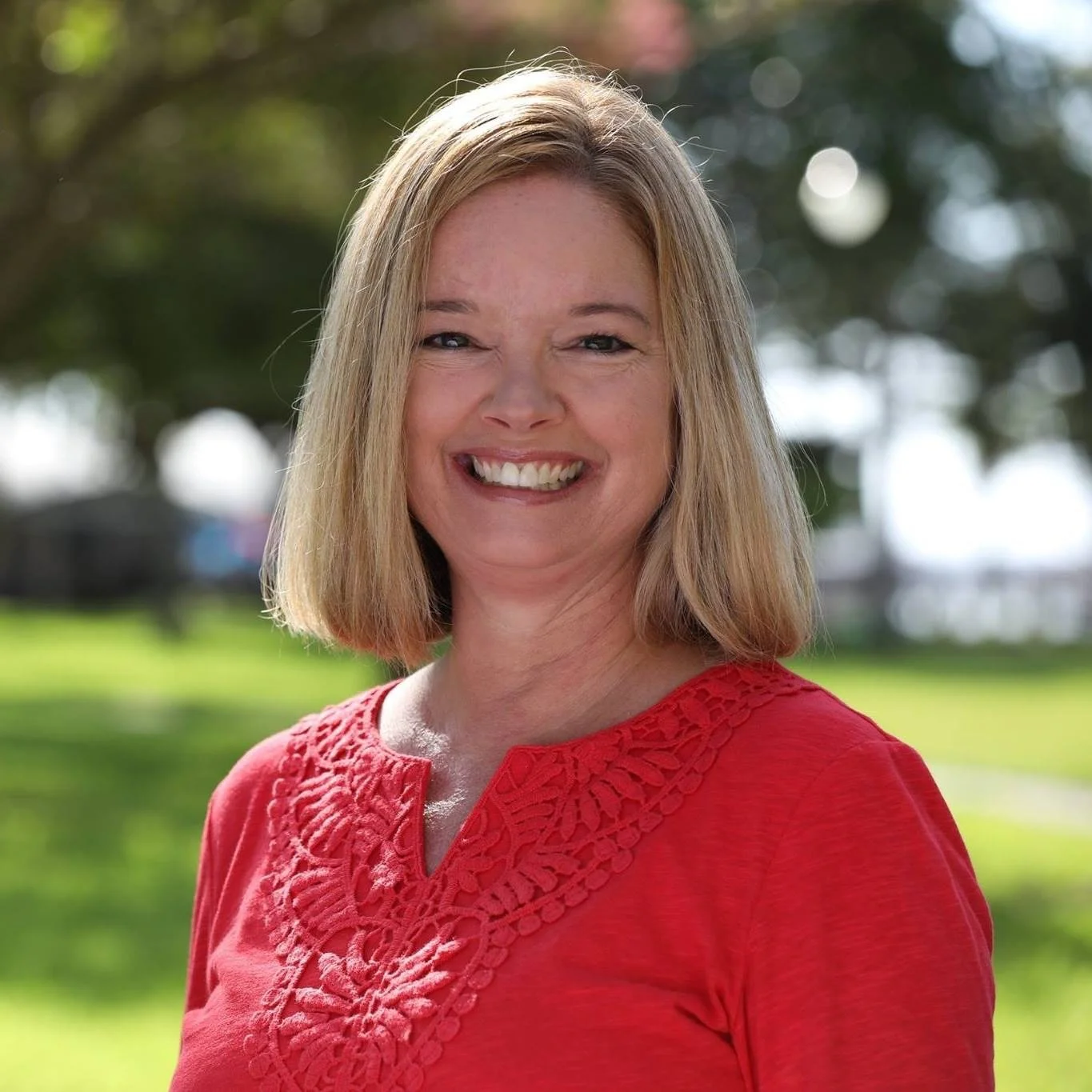 A woman with shoulder-length blonde hair, smiling, wearing a red top with embroidery details, standing outdoors with blurred green trees and bright sunlight in the background.