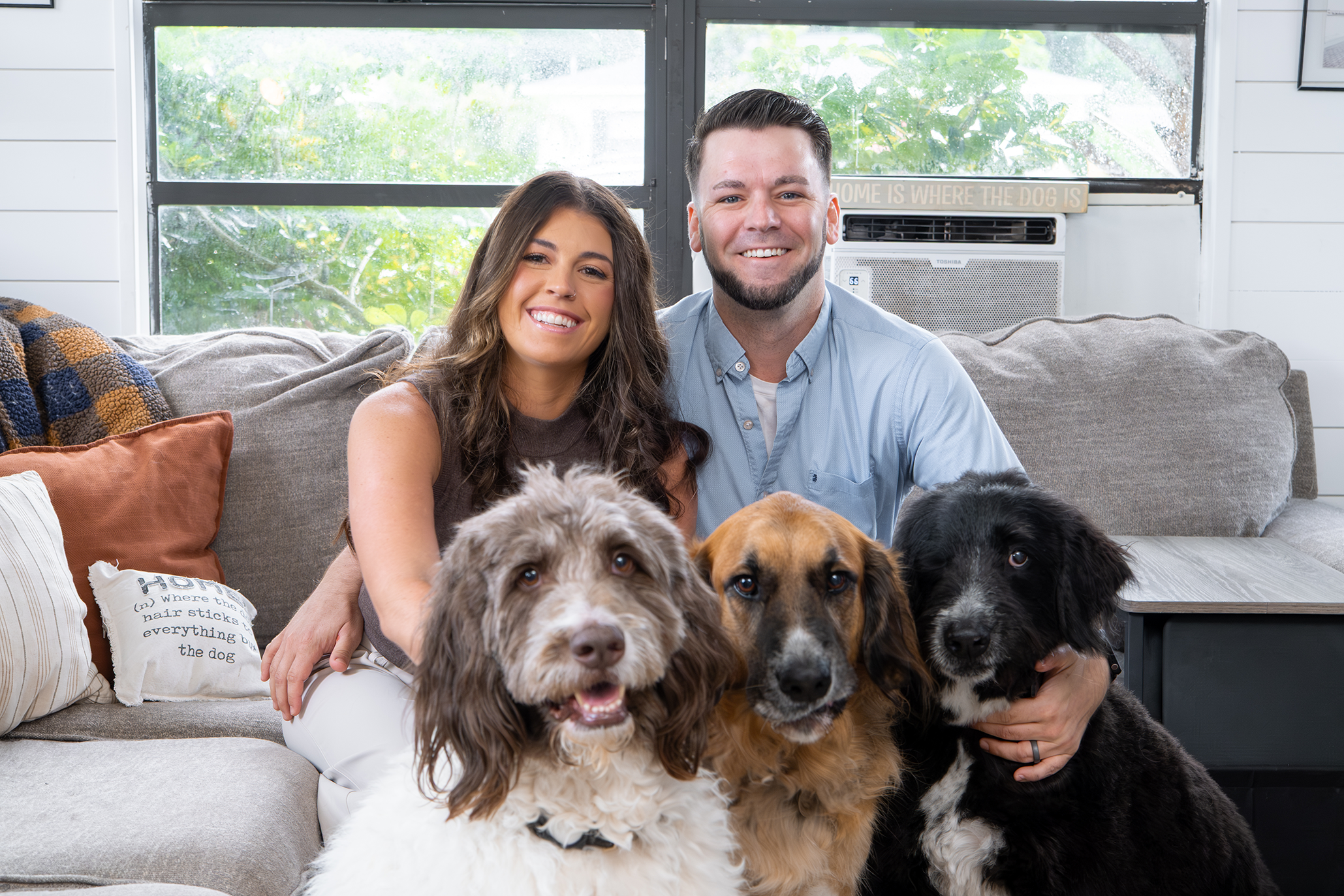 Lindsay Polega-Quigley and partner sitting on a sofa with six dogs of various breeds and colors, in a bright living room with windows in the background.