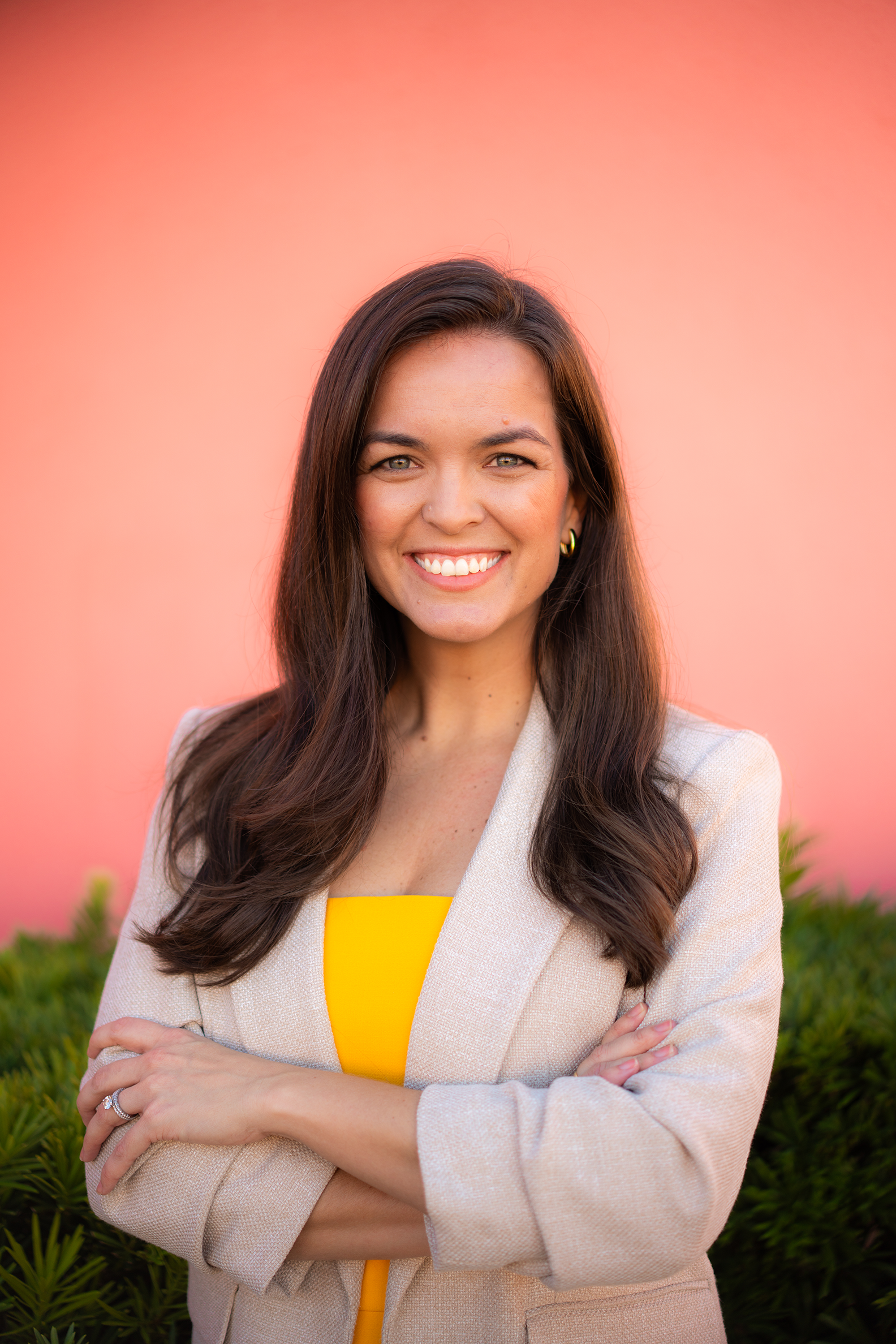 A woman smiling with arms crossed, wearing a beige blazer and a yellow top, standing outdoors in front of pink background and green bushes.