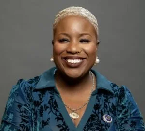 Portrait of a smiling Black woman with short blond hair wearing a blue patterned blazer, pearl earrings, and a necklace with a circular pendant, against a gray background.