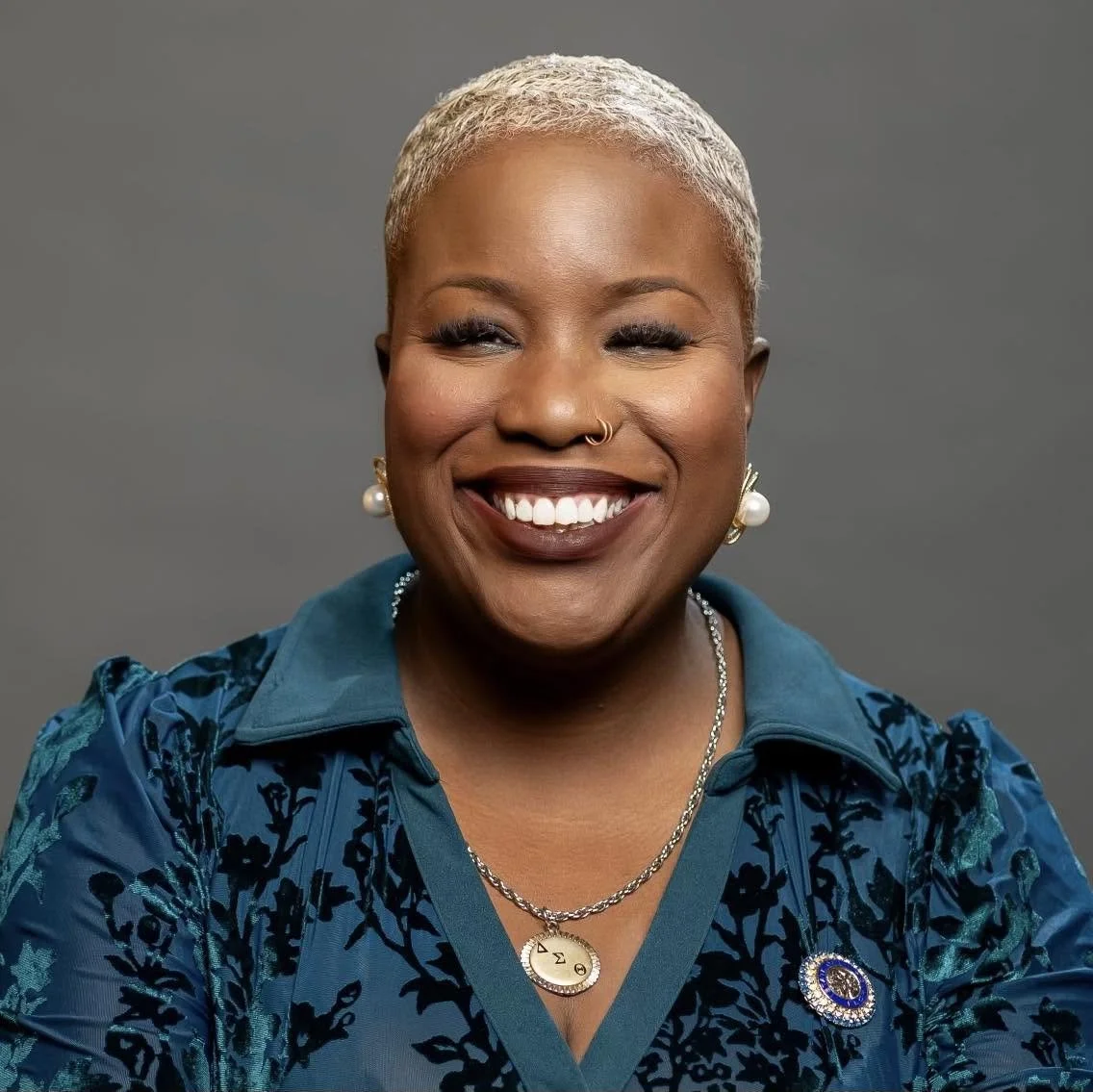 A woman with short blonde hair smiling, wearing a blue patterned blouse, pearl earrings, a necklace with an omega symbol, and a pin on her chest.