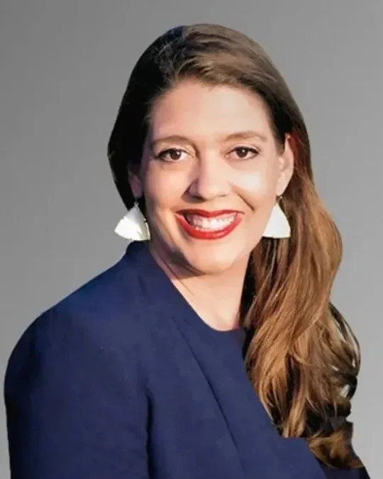 A woman with long brown hair, wearing a dark blue blazer and white earrings, smiling against a gray background.