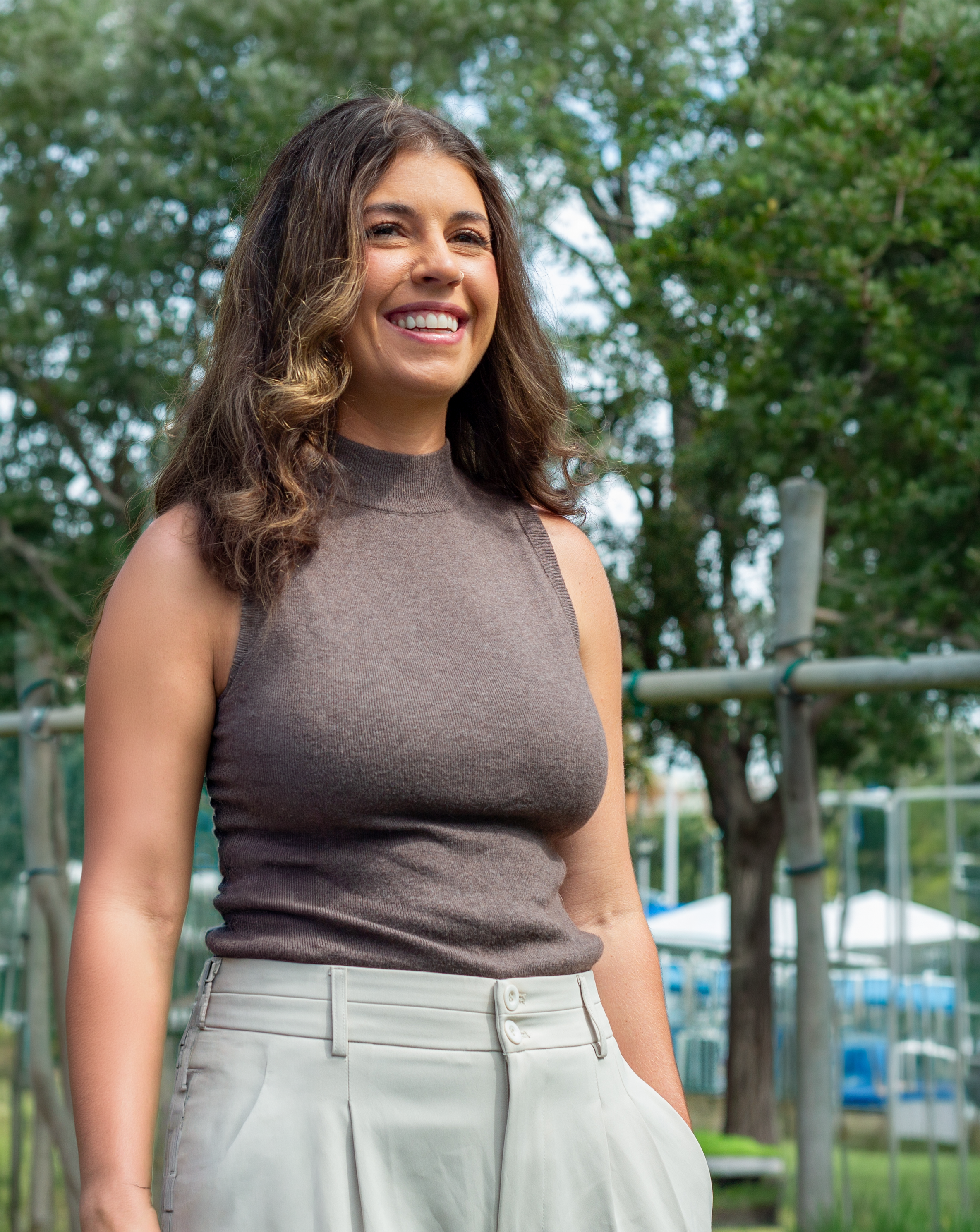 Close-up of a woman smiling outdoors, wearing a sleeveless brown top and light-colored pants, with trees and playground equipment in the background.