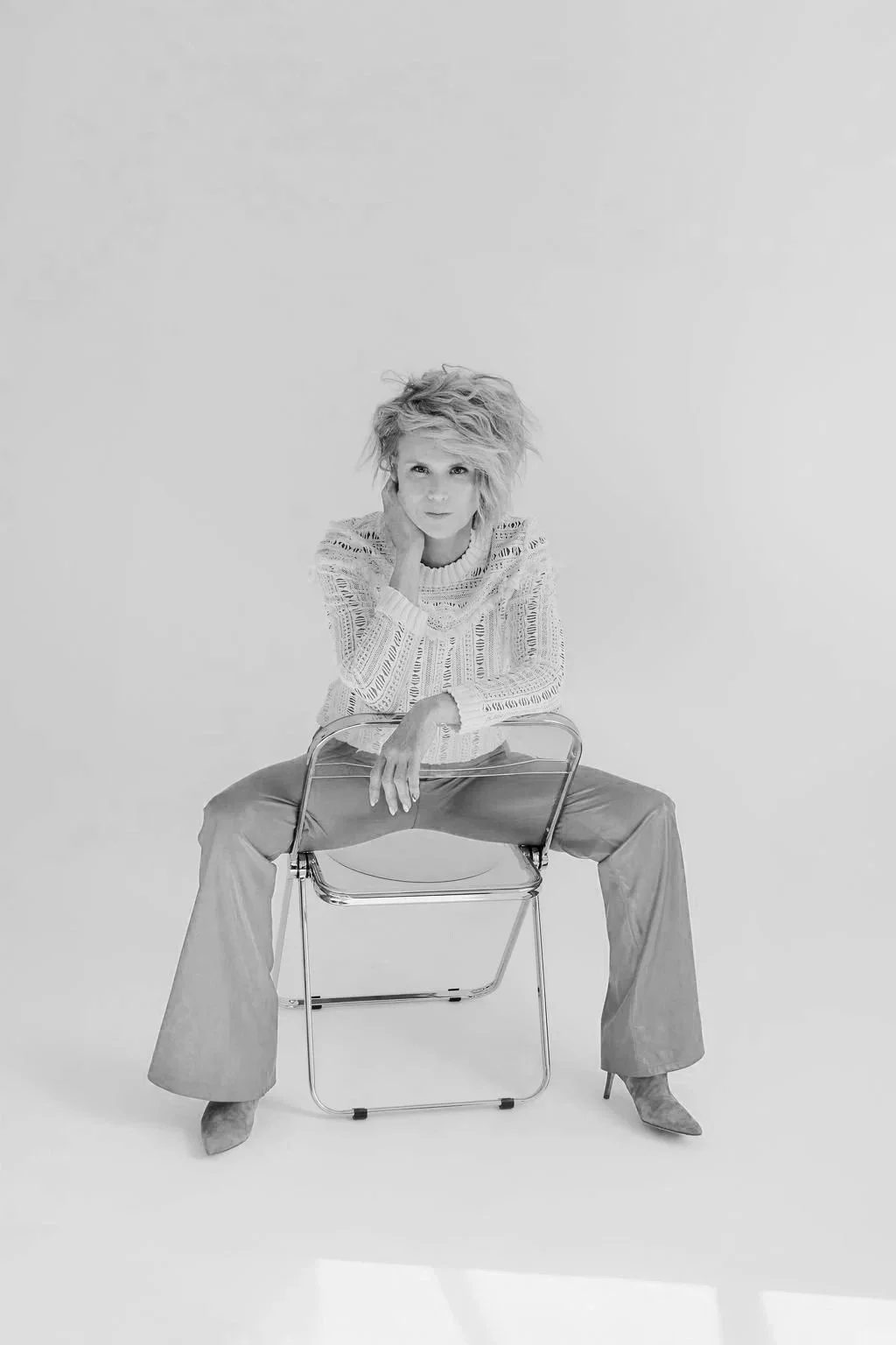 A woman with light-colored, messy hair, dressed in a patterned sweater and wide-legged pants, is sitting on a transparent chair in a studio with a plain white background. She rests her head on her left hand and looks at the camera.