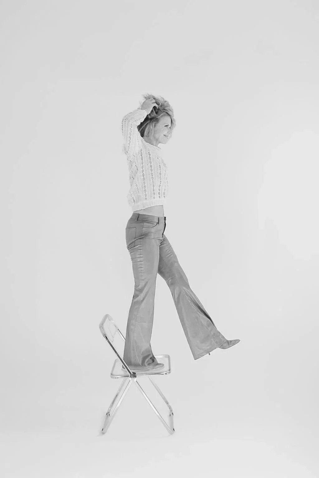 A woman balancing on a transparent chair with one foot, smiling with her hand in her hair, wearing a knit sweater and flared pants, in a white studio setting.