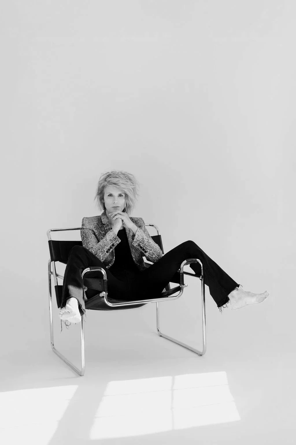 Black and white photo of a woman sitting on a modern chair, with her legs crossed and hands resting under her chin, against a plain background.